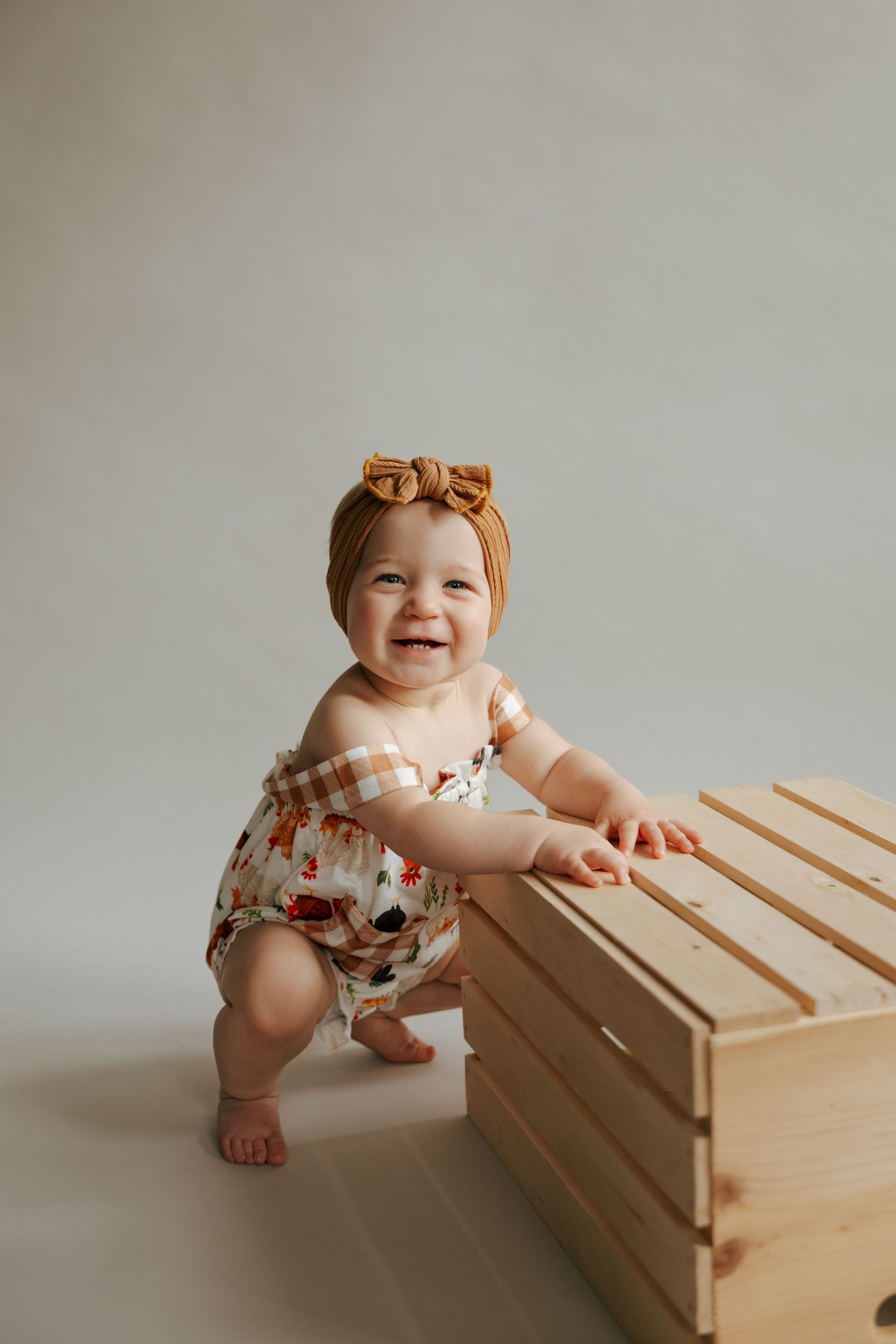 Happy baby girl in floral romper and mustard headband standing beside a wooden crate.
