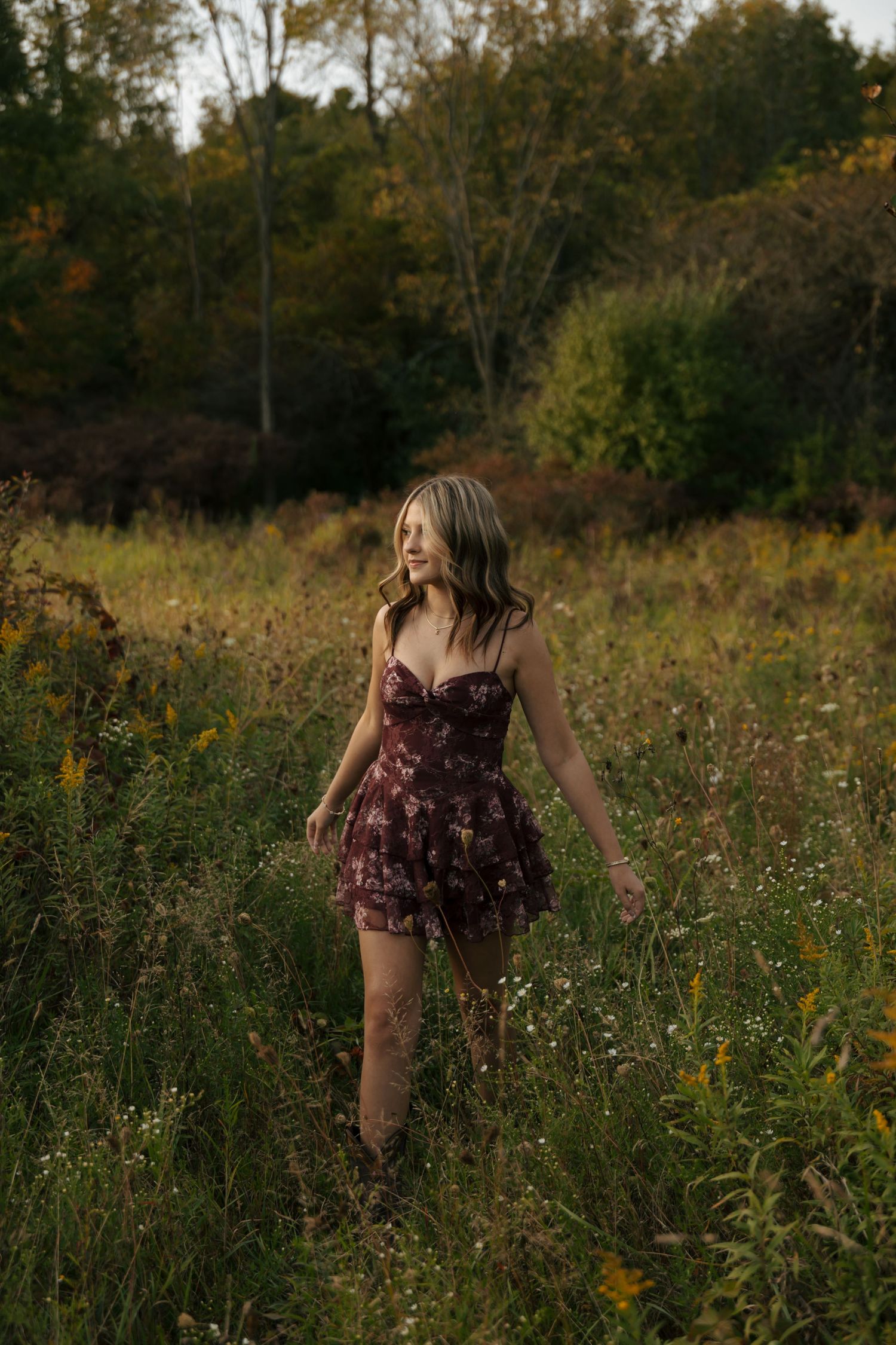 Young woman in burgundy floral mini dress walking through autumn wildflower meadow with colorful fall foliage background.