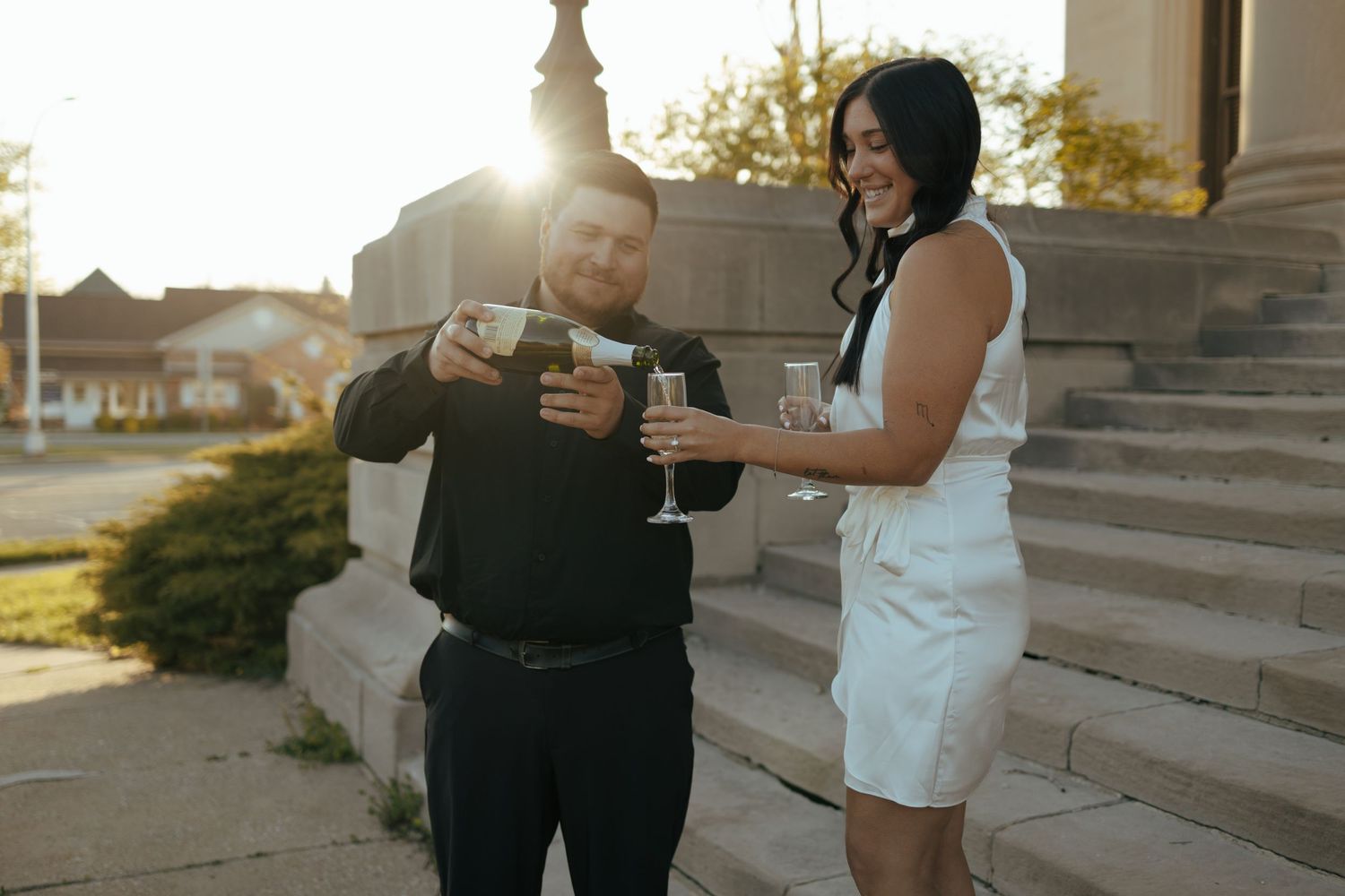 Elegant couple toasting champagne glasses outdoors on steps bathed in warm golden sunset light.