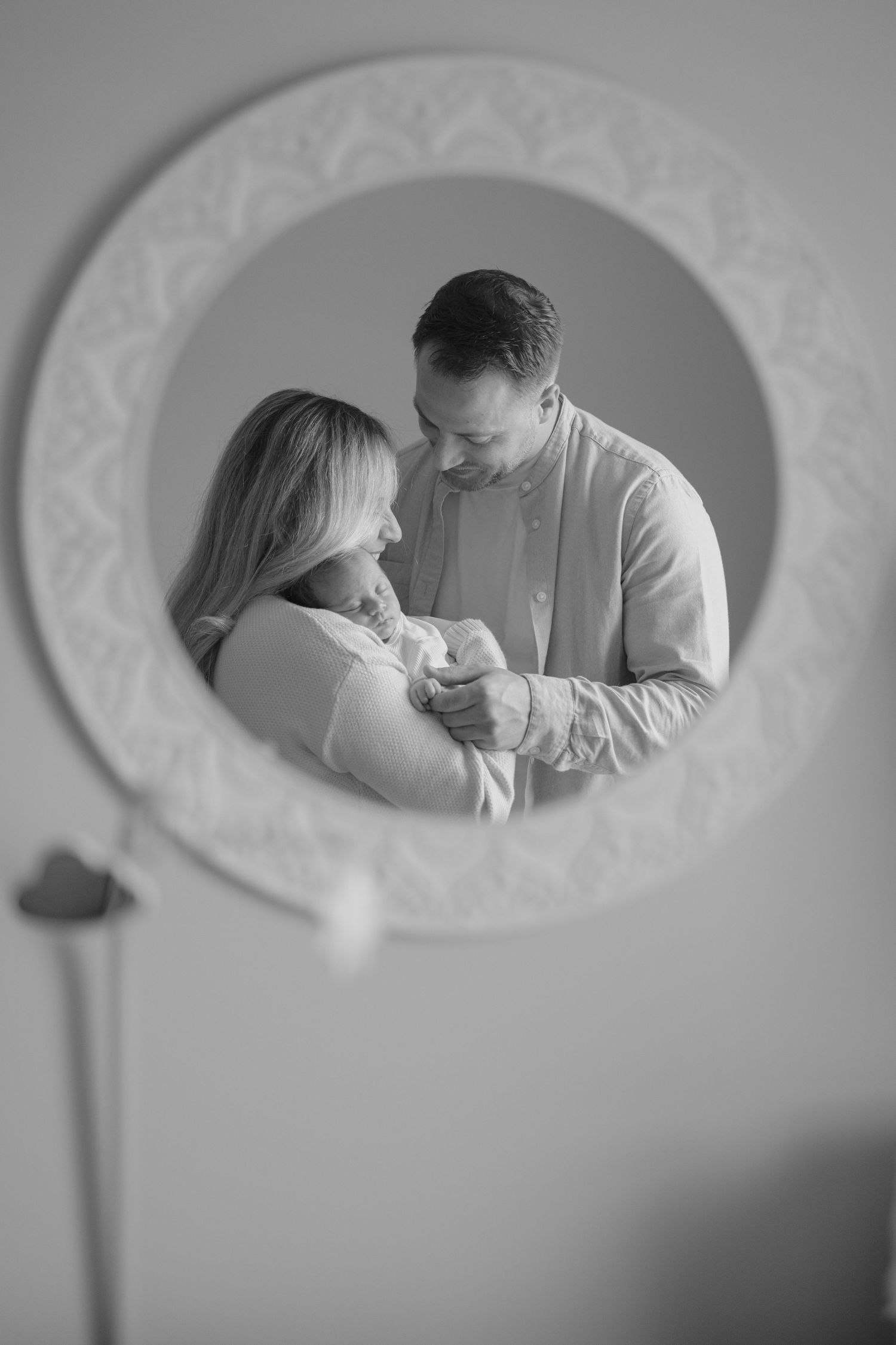 Black and white reflection of new parents lovingly holding their newborn baby in round mirror.