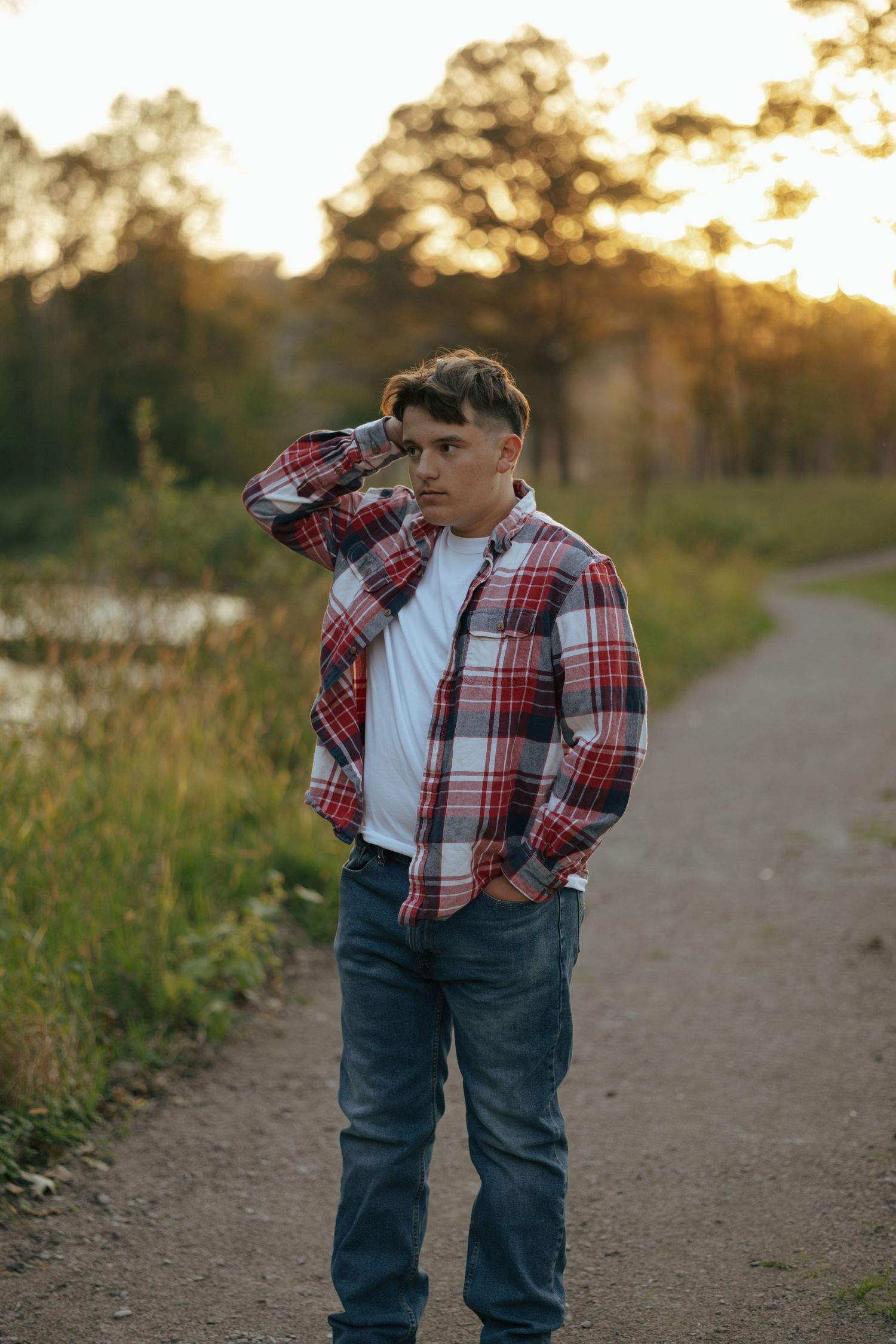 Young man in red plaid flannel shirt stands on park path at golden hour near pond.
