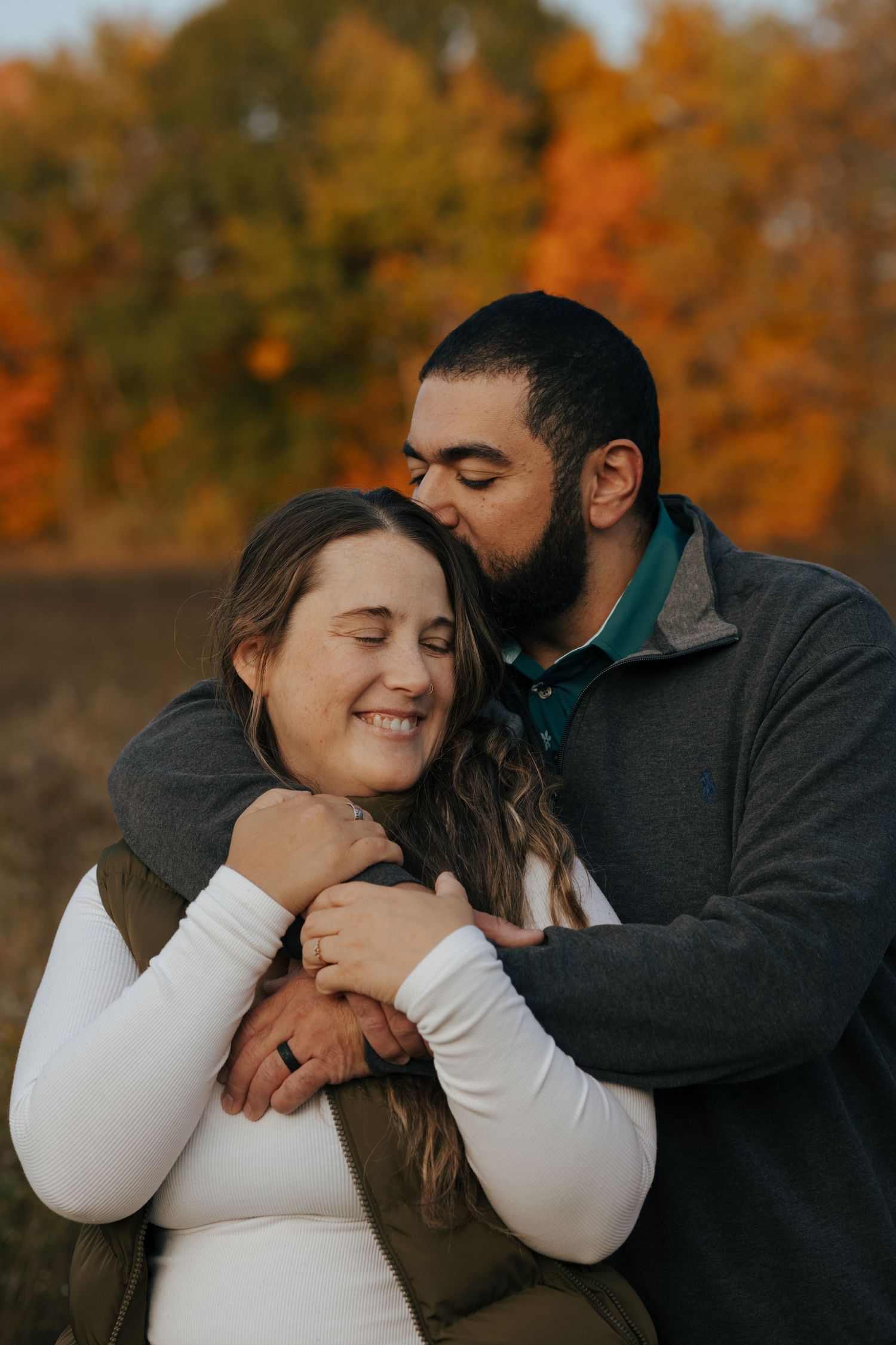 Bearded man embracing and kissing smiling woman with colorful fall foliage in background.