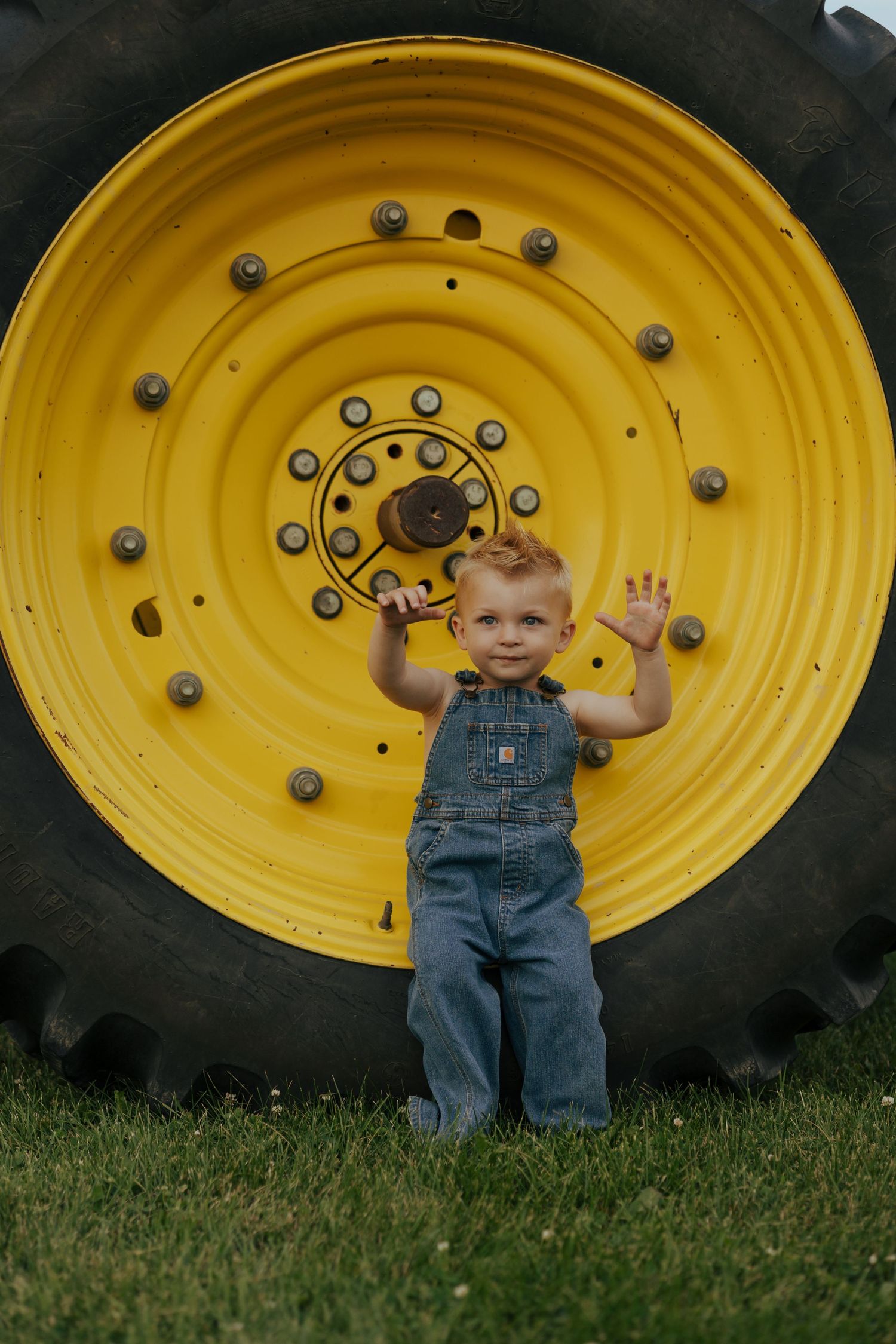 Happy toddler in denim overalls standing in front of large yellow tractor tire outdoors.