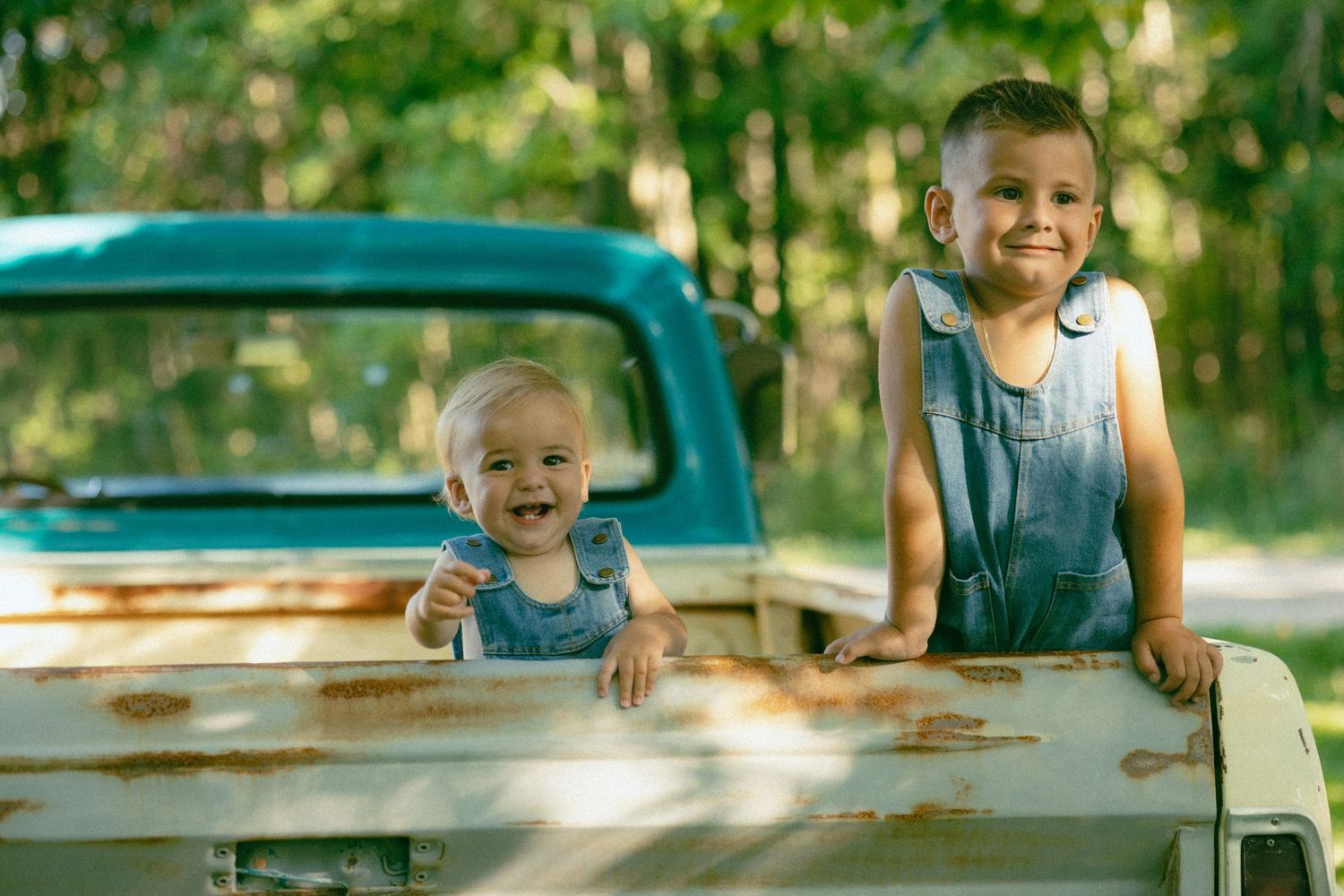 Two young siblings in matching denim overalls smiling in bed of vintage teal pickup truck.