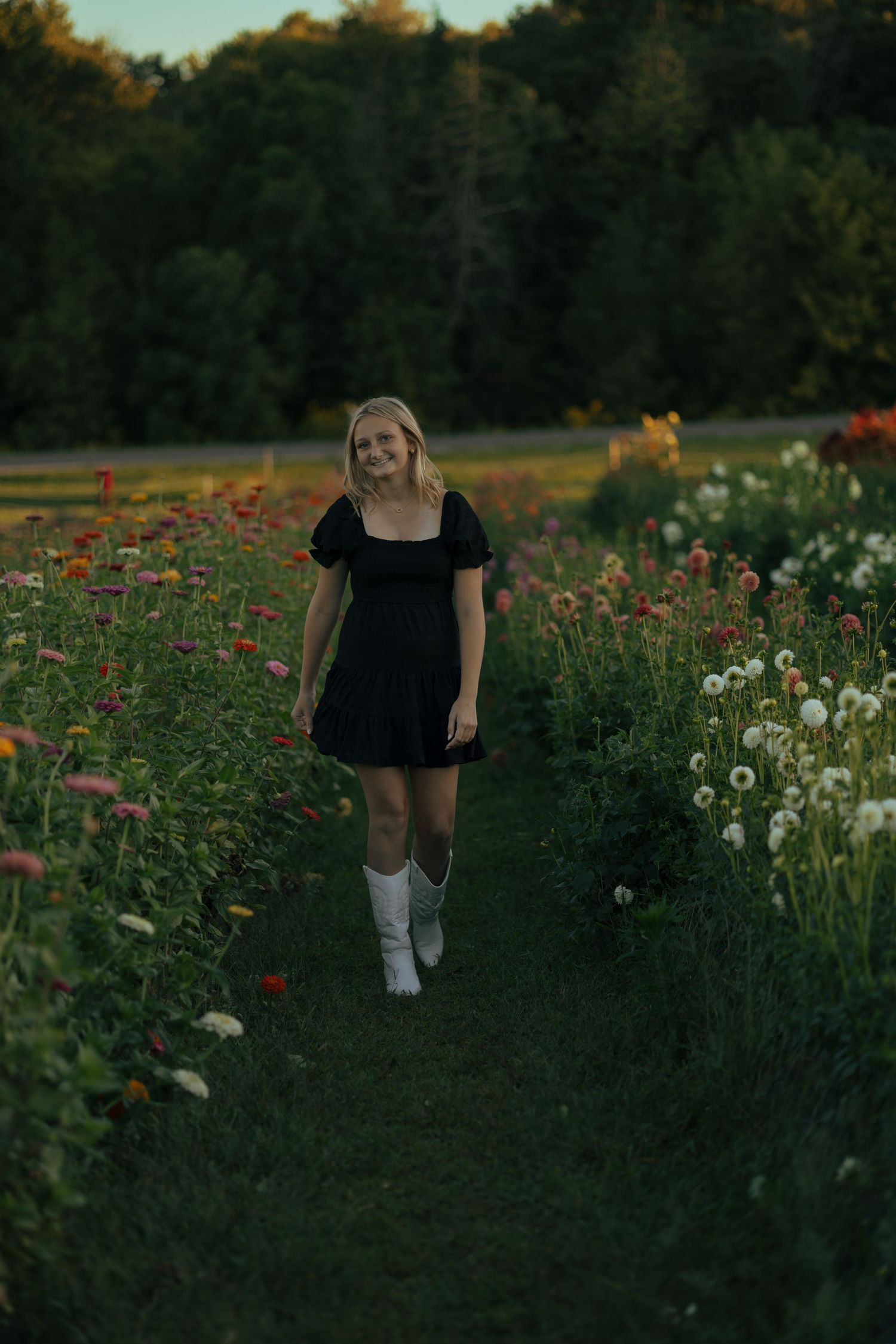 Blonde teen girl in black mini dress and white boots walking through colorful flower garden.