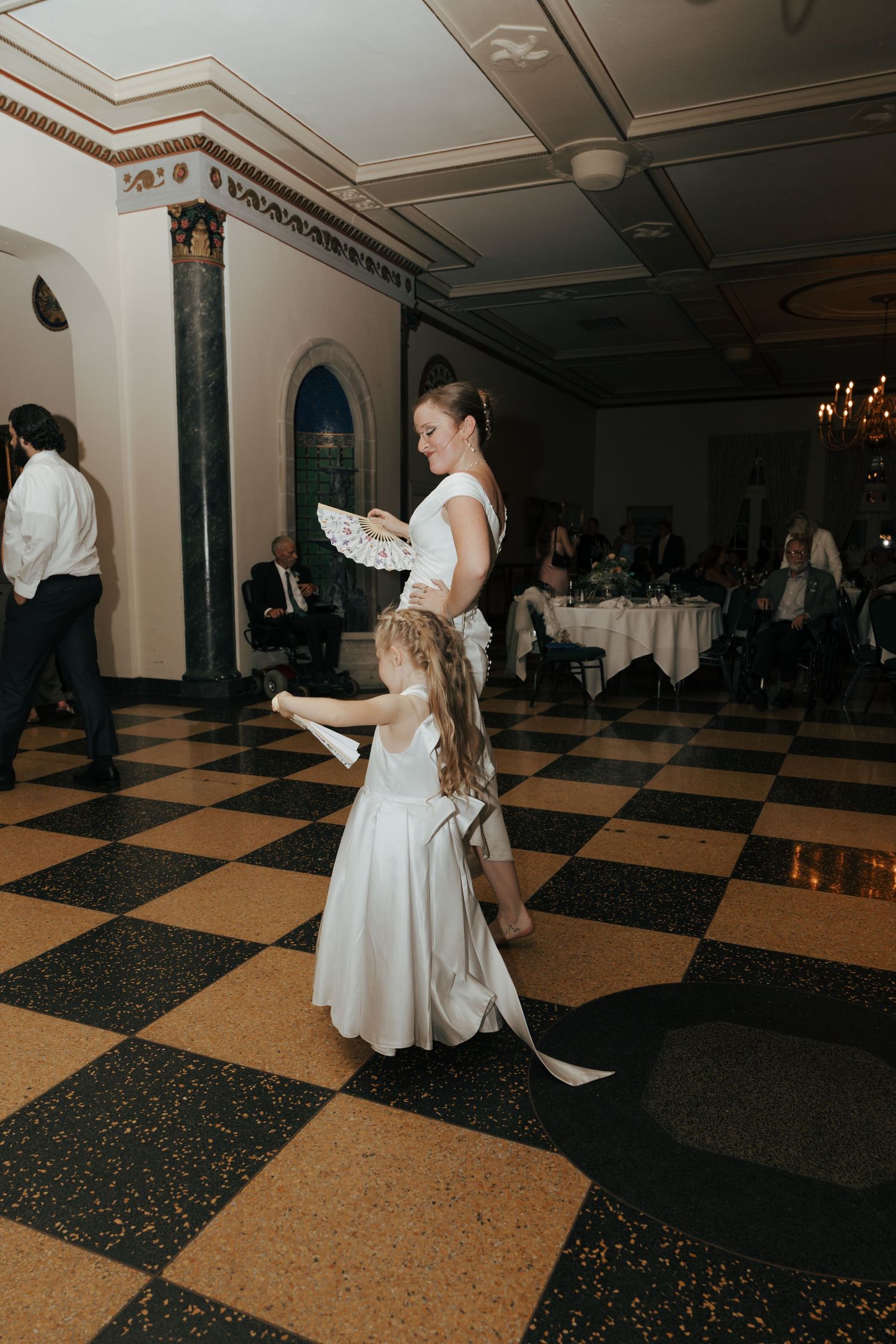 Bride and flower girl dancing together holding decorative fans on checkered floor.
