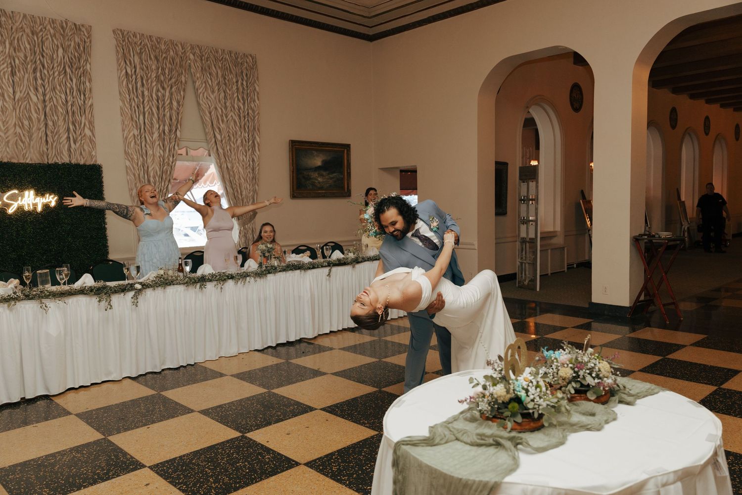 Groom dipping bride dramatically on checkered dance floor at wedding reception with guests watching.