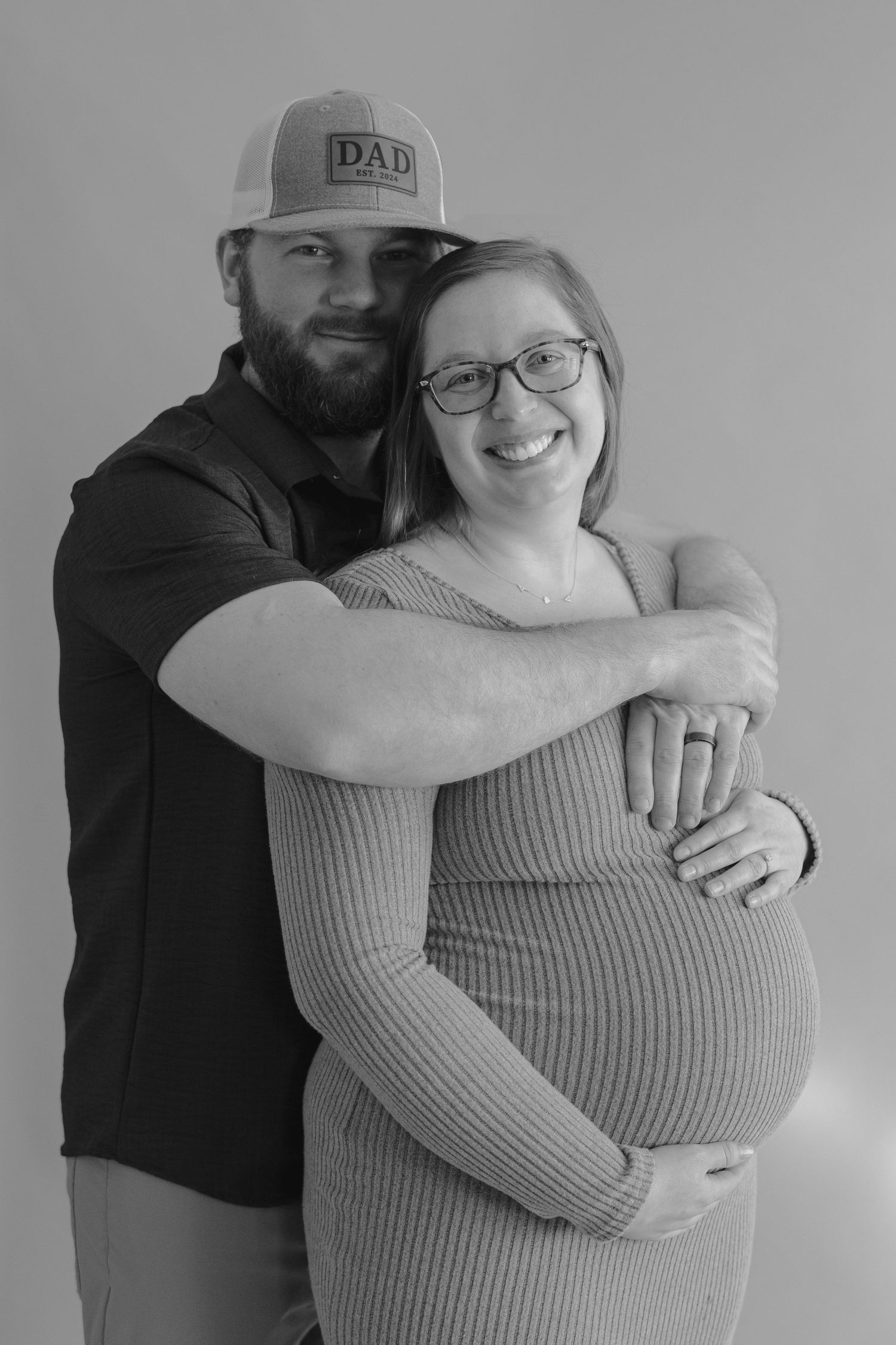 Black and white photo of smiling pregnant woman in ribbed dress embraced by bearded man in DAD cap.
