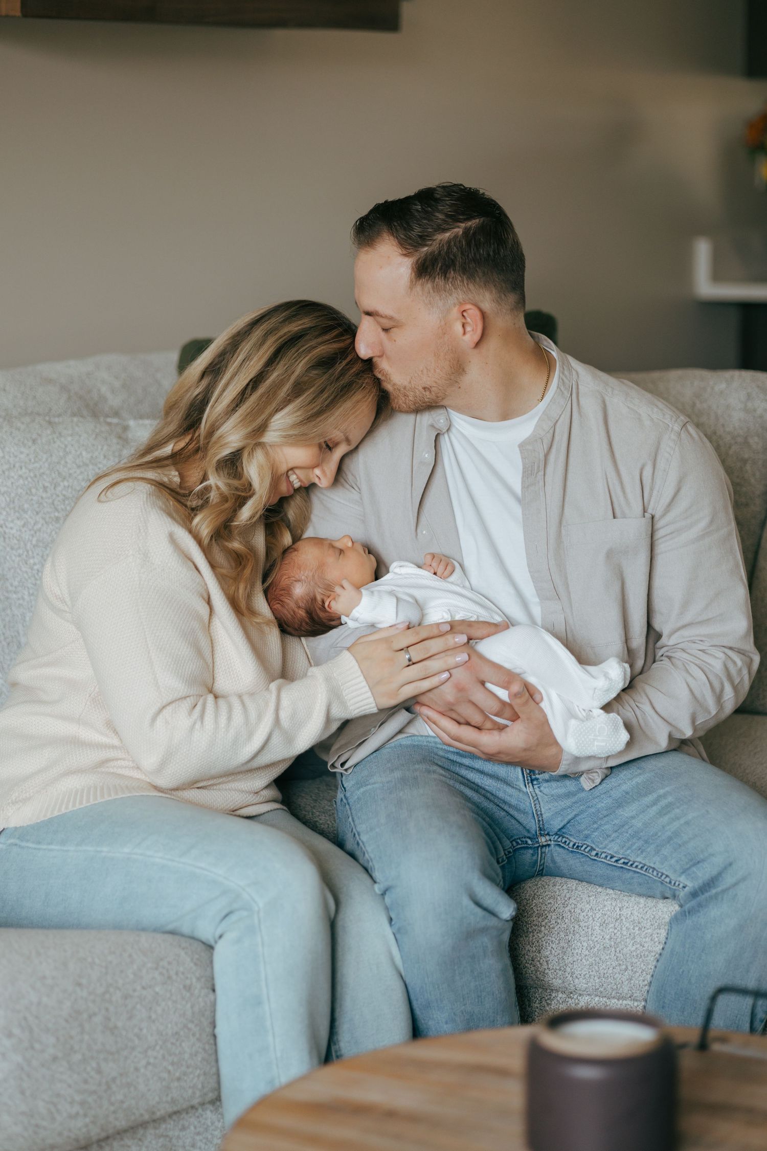 New parents sit on couch smiling as father holds swaddled newborn baby in cozy home setting.