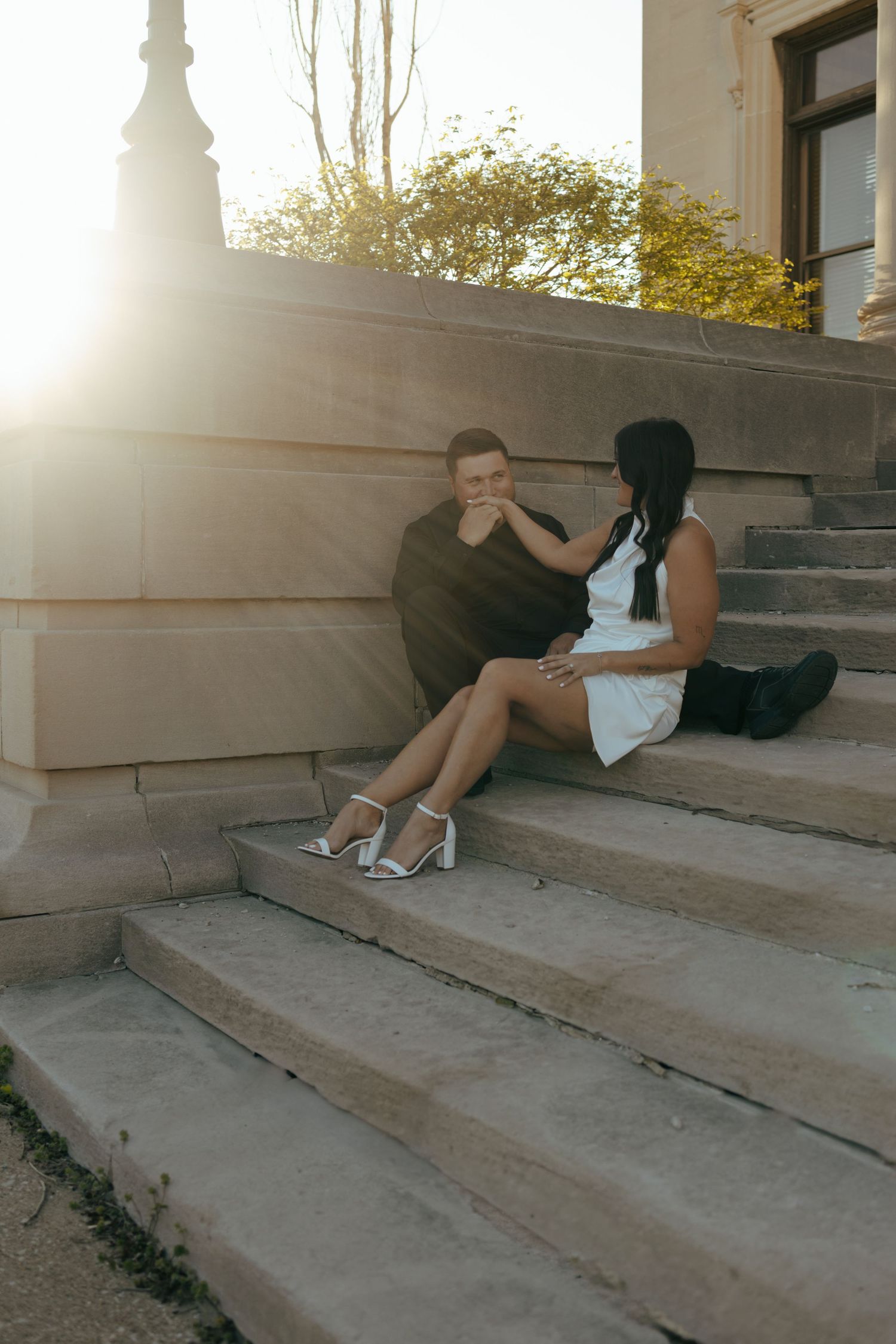 Couple sitting on stone steps at sunset, man kissing woman's hand in romantic moment.