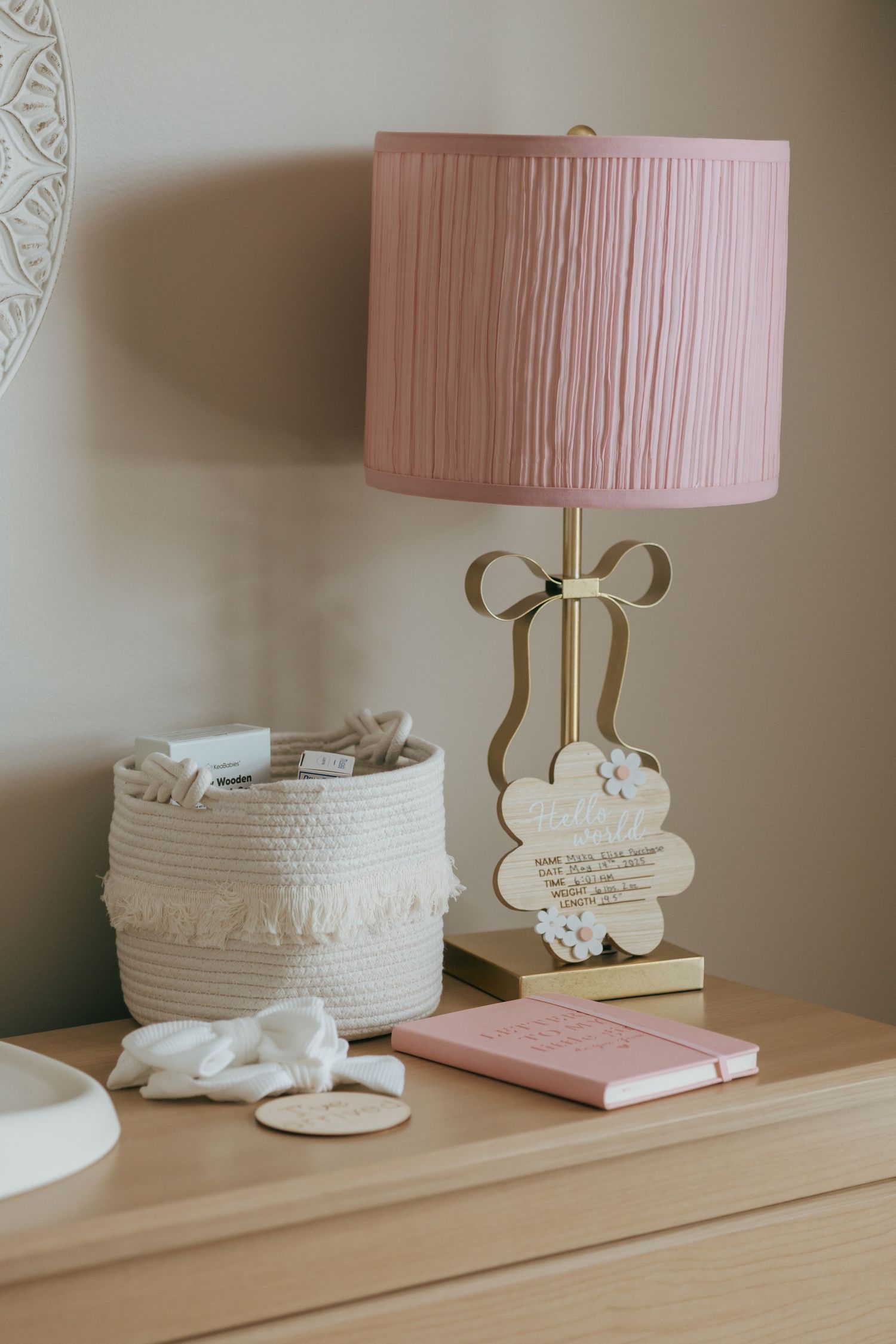Pink pleated lamp with gold bow base beside a woven basket and pink journal on a dresser.