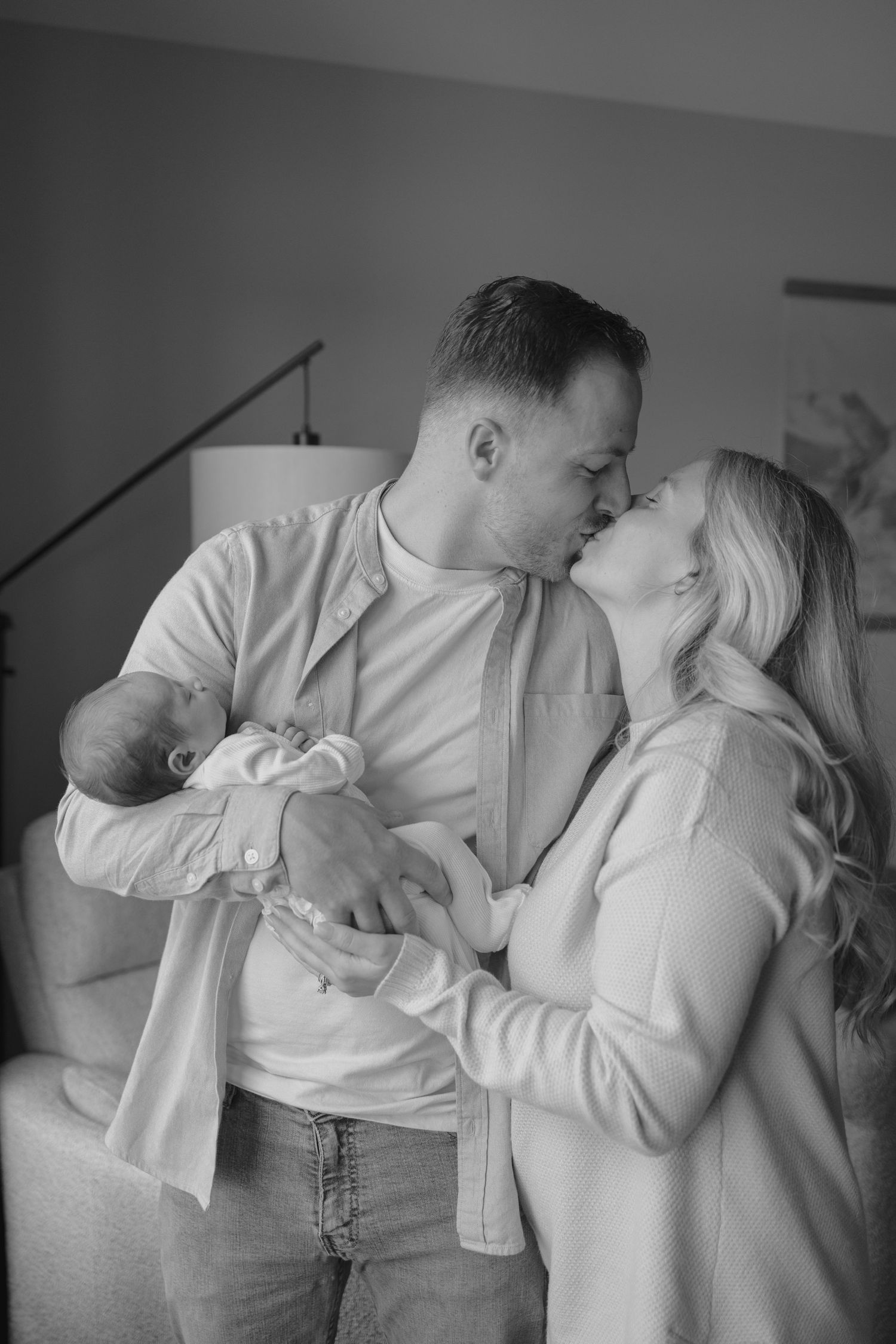 Black and white photo of couple kissing while tenderly holding their newborn baby indoors.