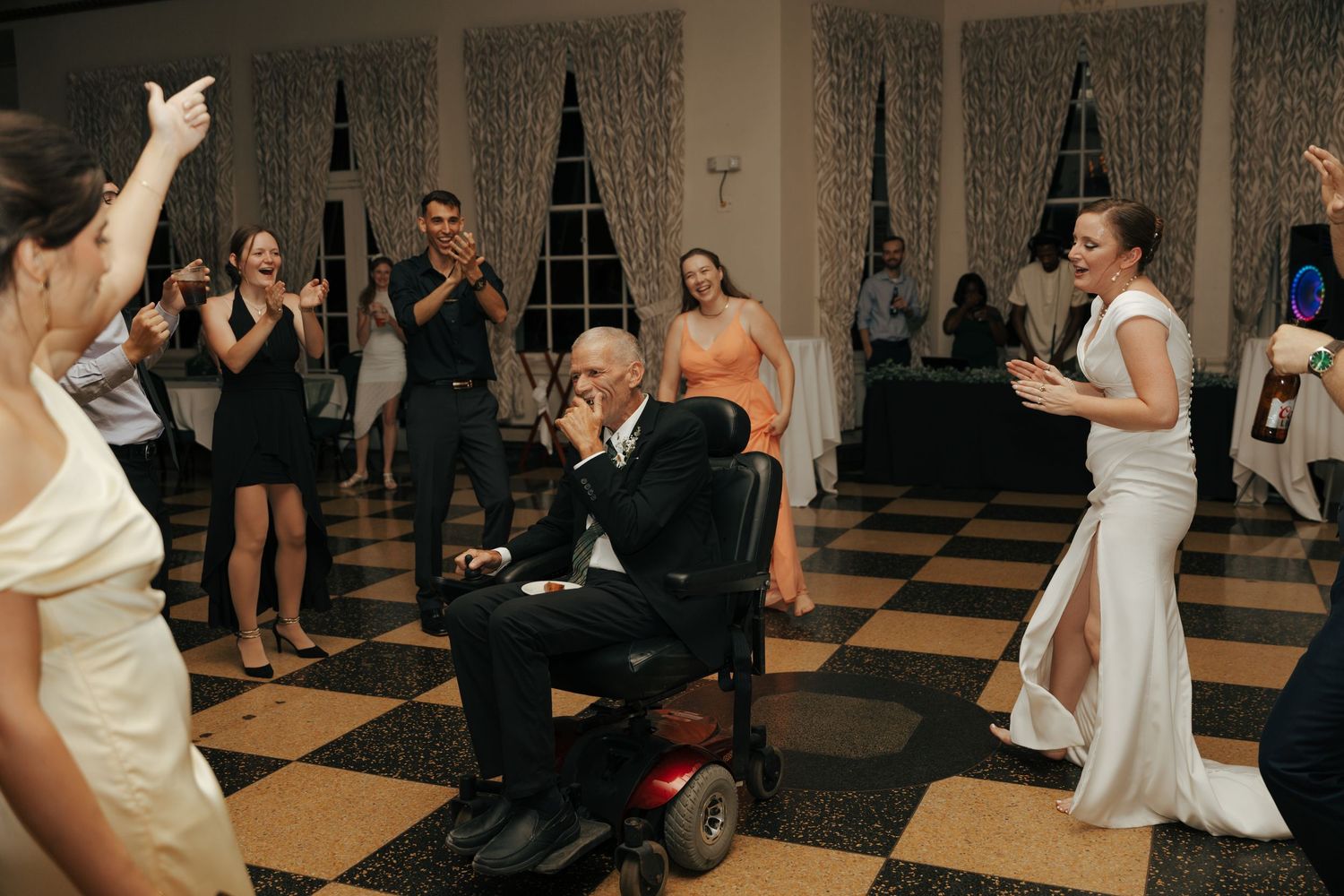 Wedding guests celebrating with elderly man in wheelchair on checkered dance floor.