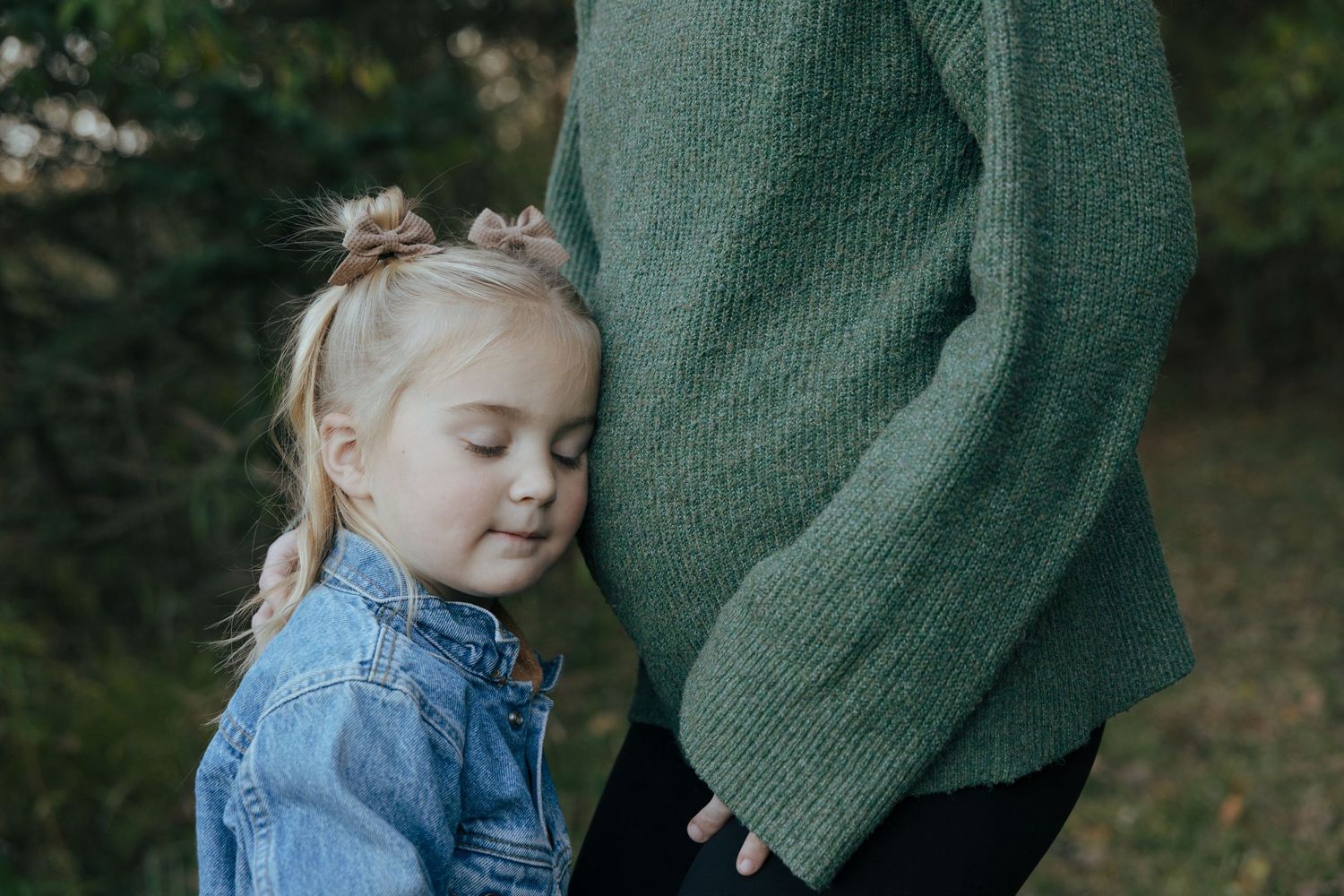 Sweet little girl in denim jacket resting her head against her pregnant mothers green sweater outdoors.