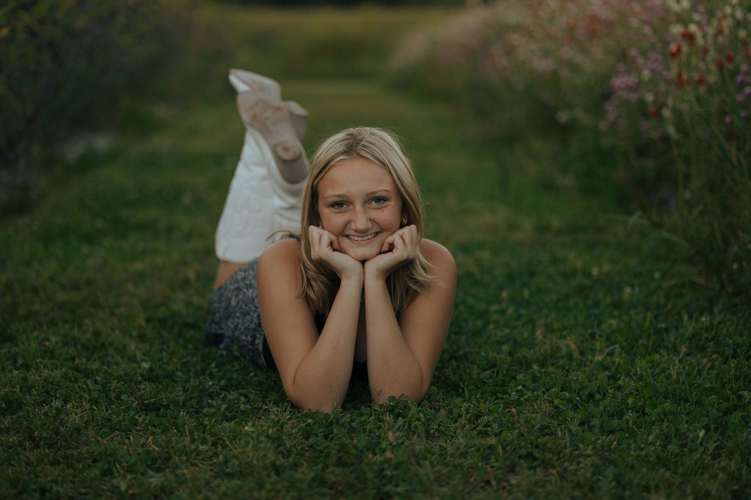 Smiling blonde teenage girl lying on green grass with hands under chin in garden path.