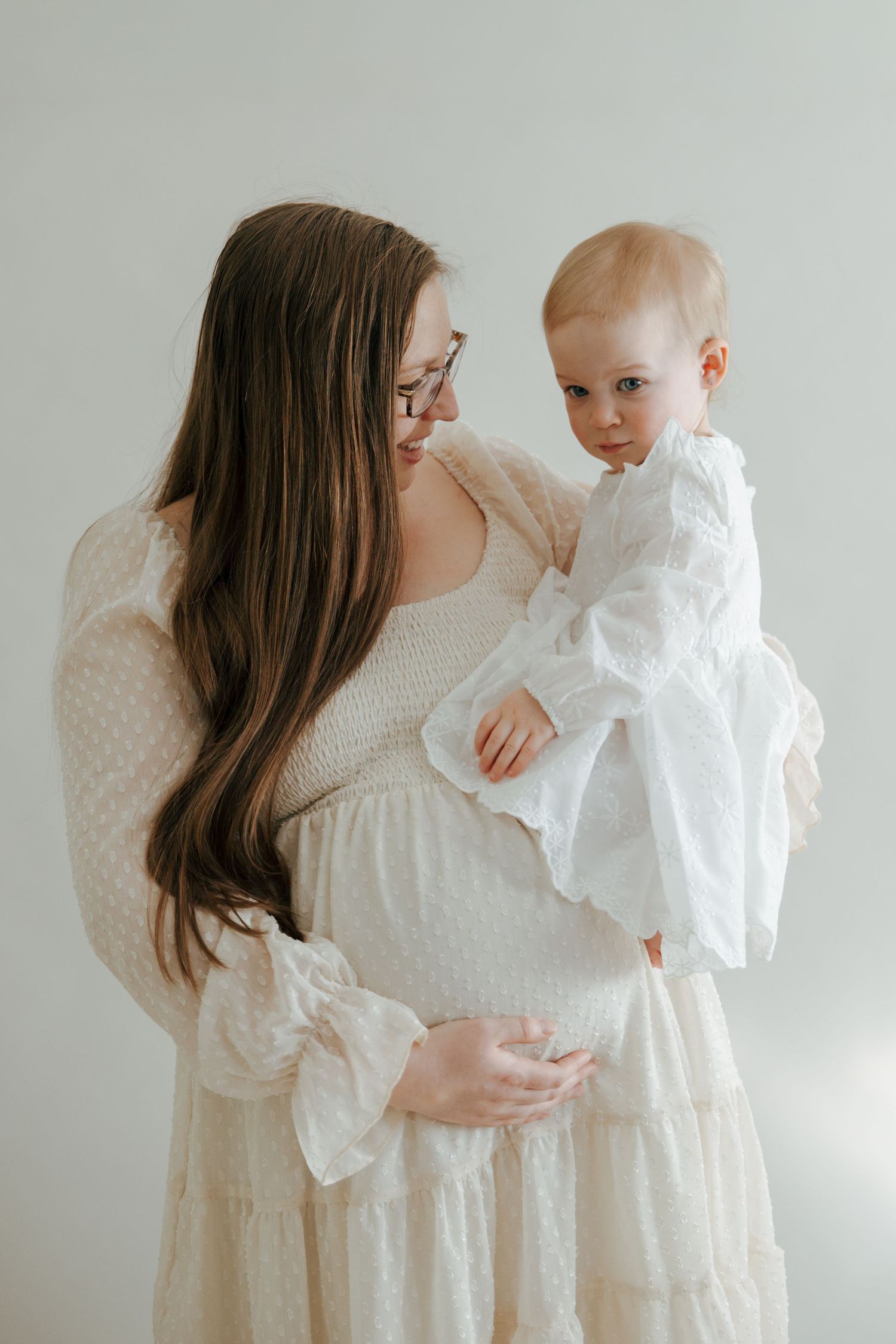 Pregnant woman in cream dress holding baby girl in white outfit against light background.