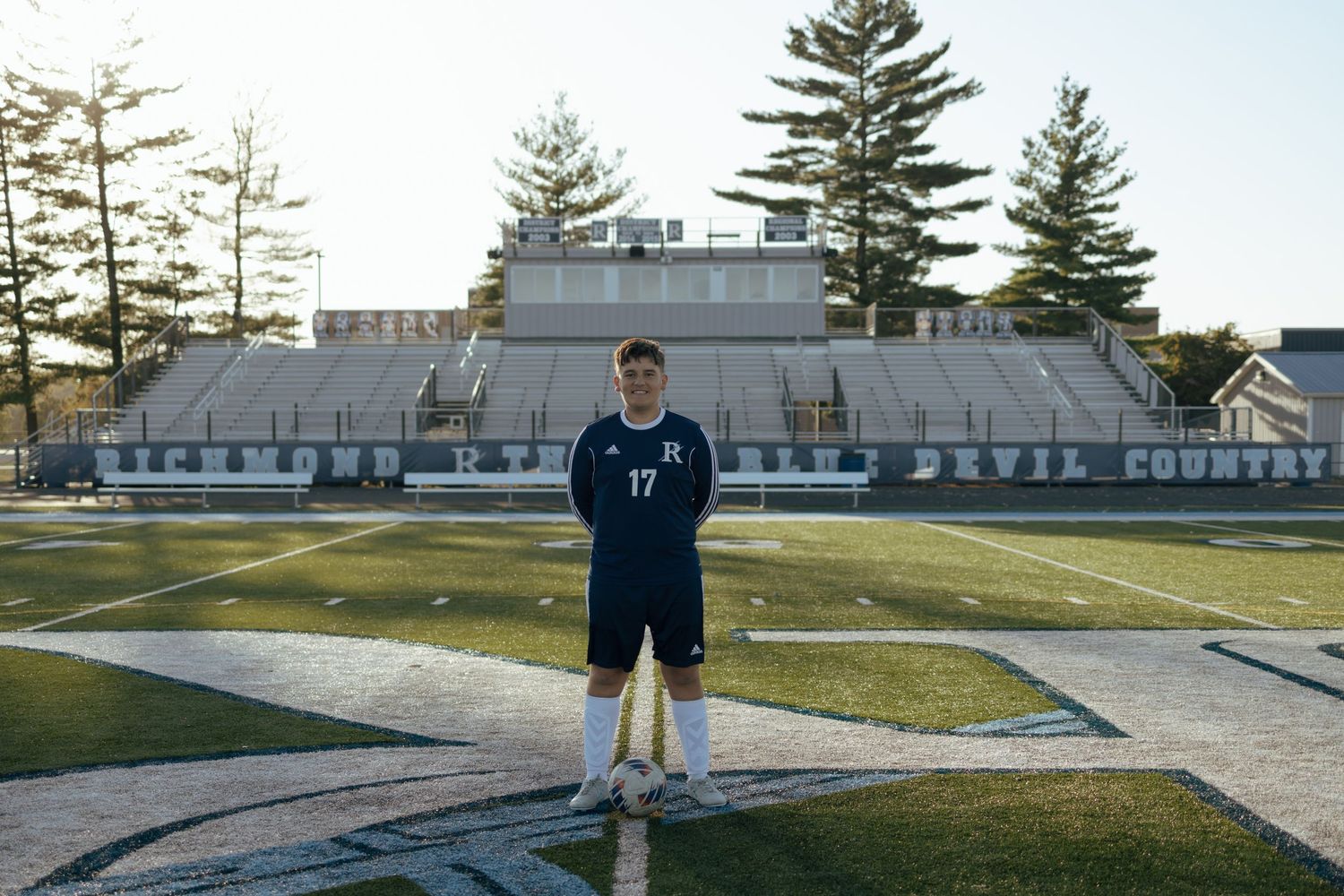 Teen soccer player #17 stands centered on Richmond Blue Devils turf field at sunset.