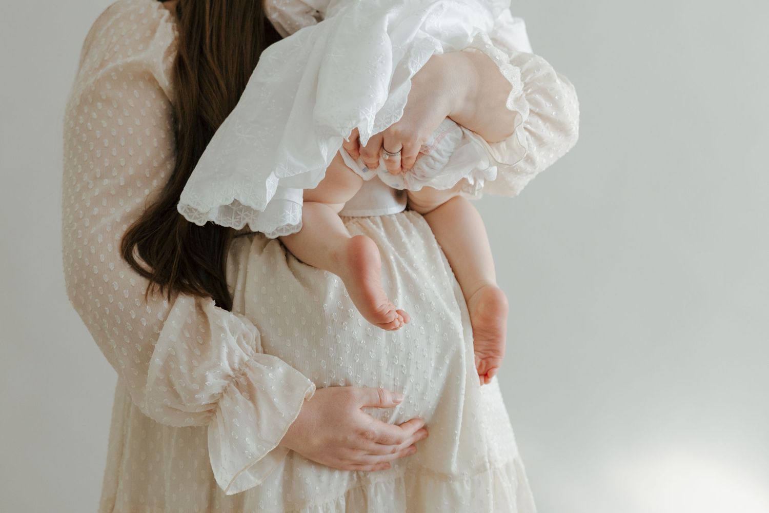 Pregnant woman in cream dotted dress gently holding sleeping baby against her bump.