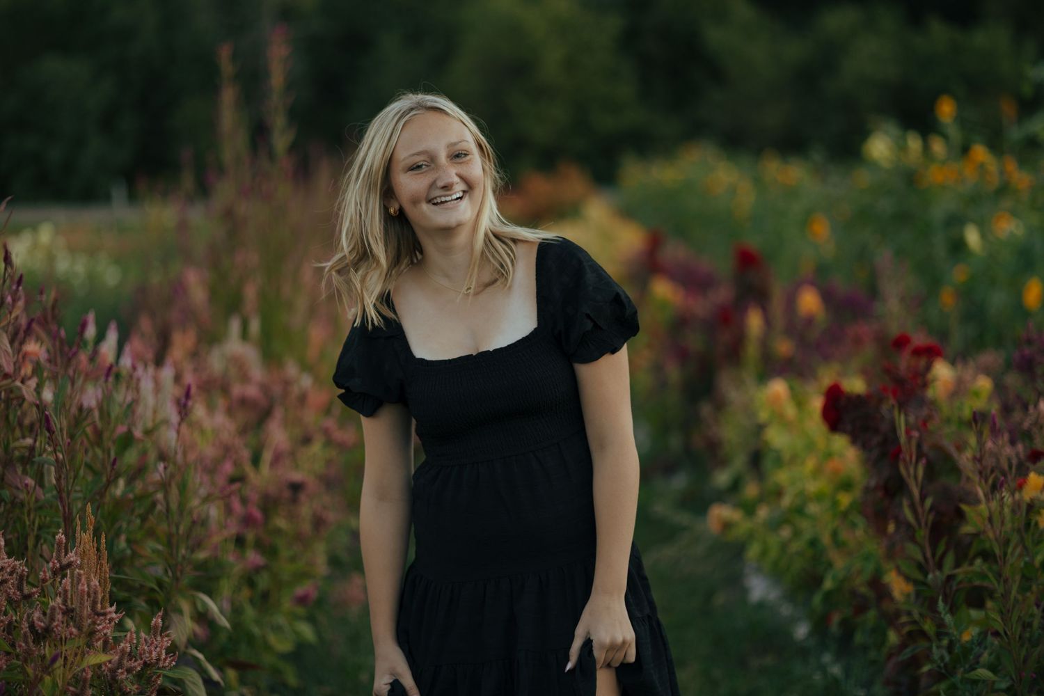 Laughing blonde young woman in black dress surrounded by colorful garden flowers outdoors.