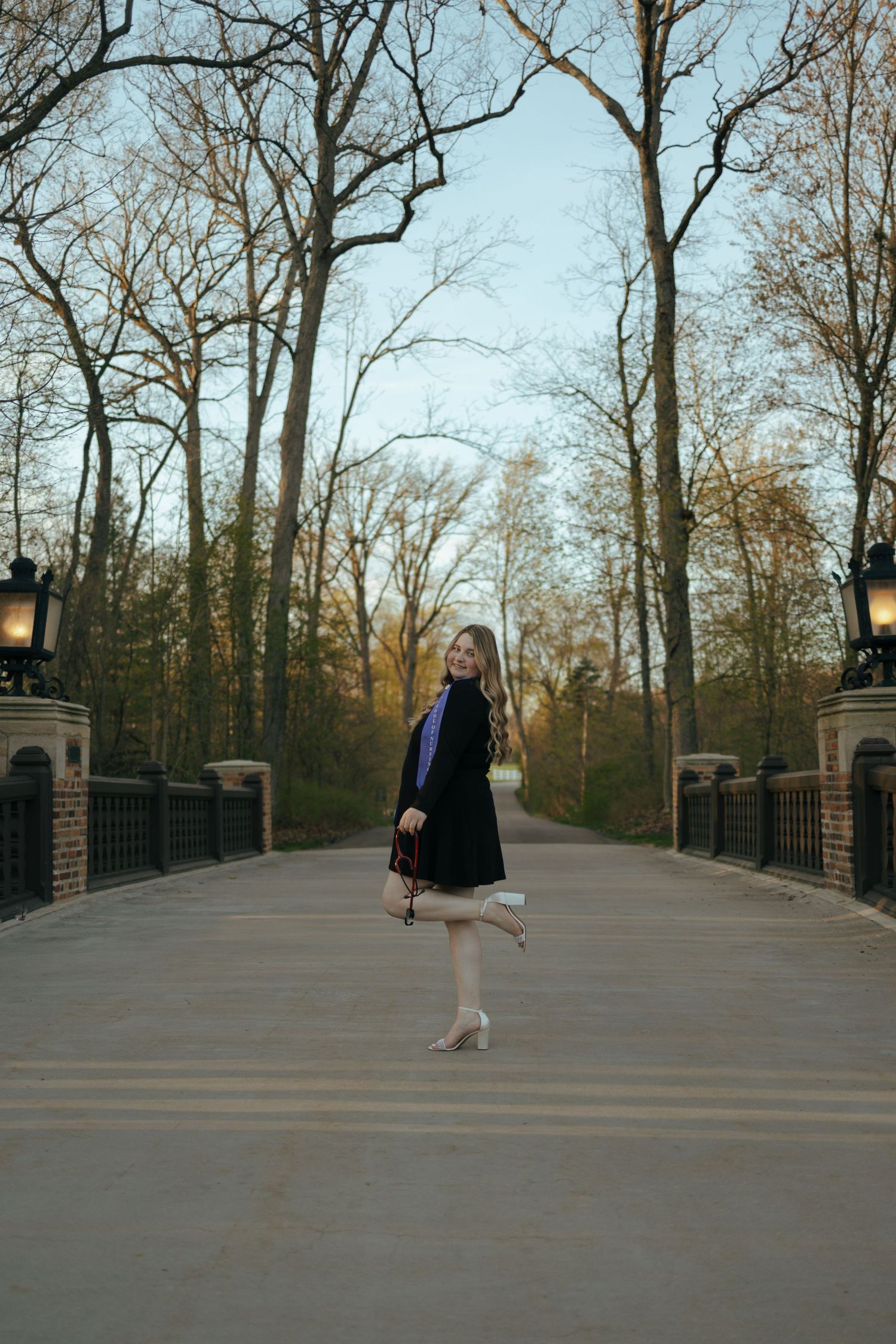 Young woman graduate poses playfully on a bridge surrounded by bare trees at dusk.