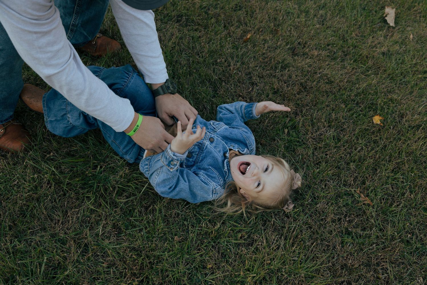 Laughing little girl lying on green grass being tickled by parent during outdoor family photo session.