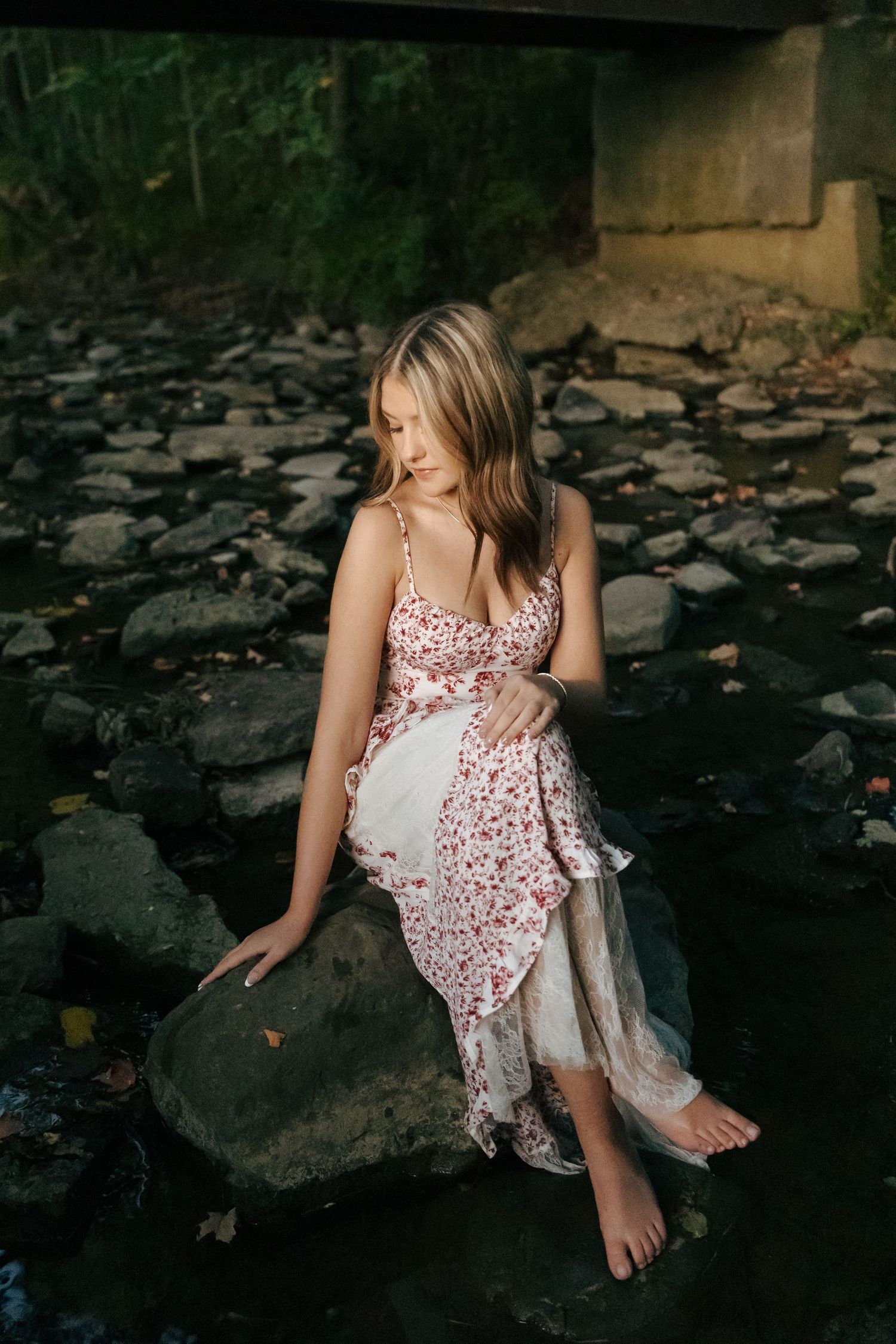Young woman in floral dress sitting on a rock in a creek beneath a rustic bridge.