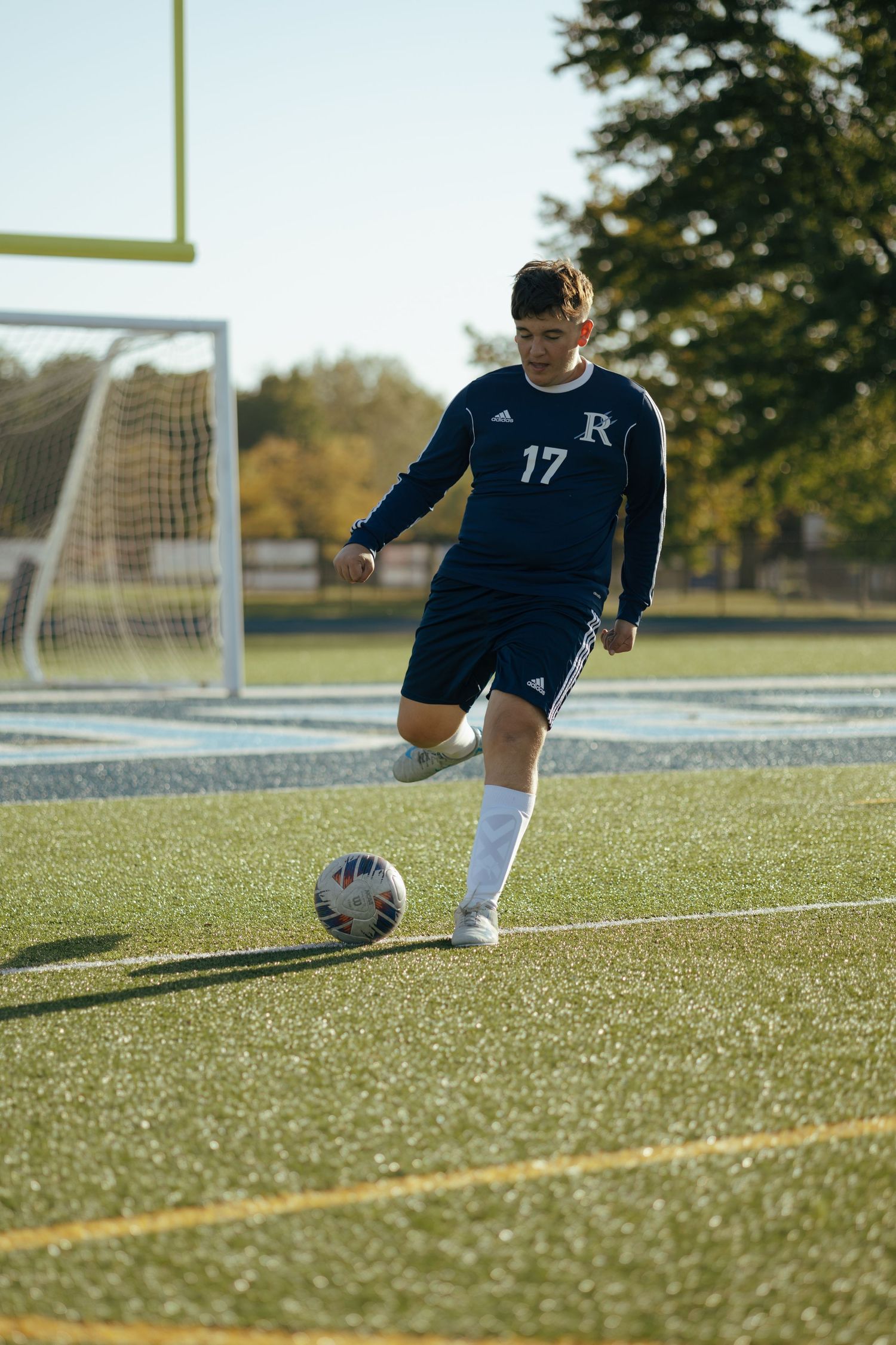 High school soccer player #17 in navy Adidas kit prepares to kick ball on turf field.
