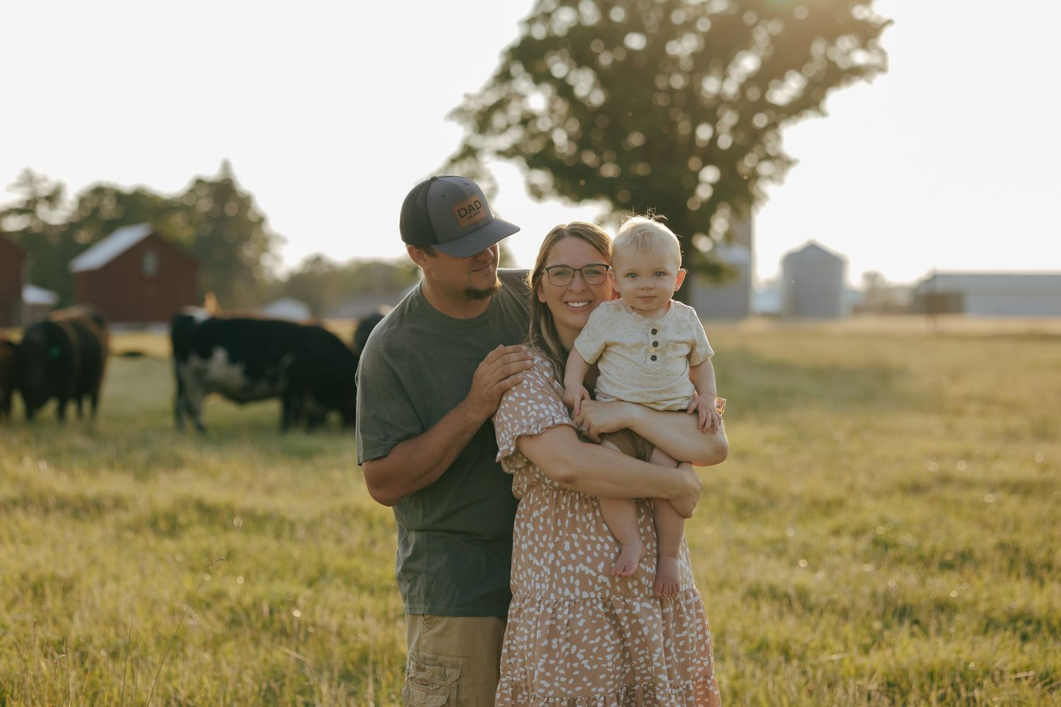 Happy young family with baby posing in farm field with cattle grazing in background.