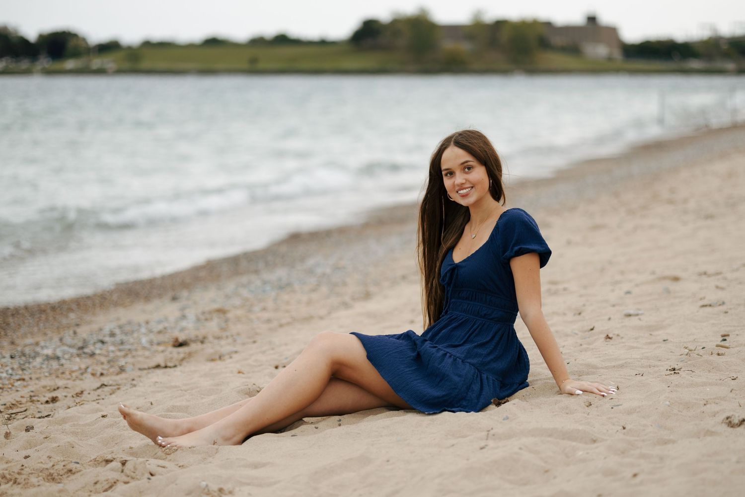 Young woman in navy blue dress sitting barefoot on sandy beach shore near calm water.