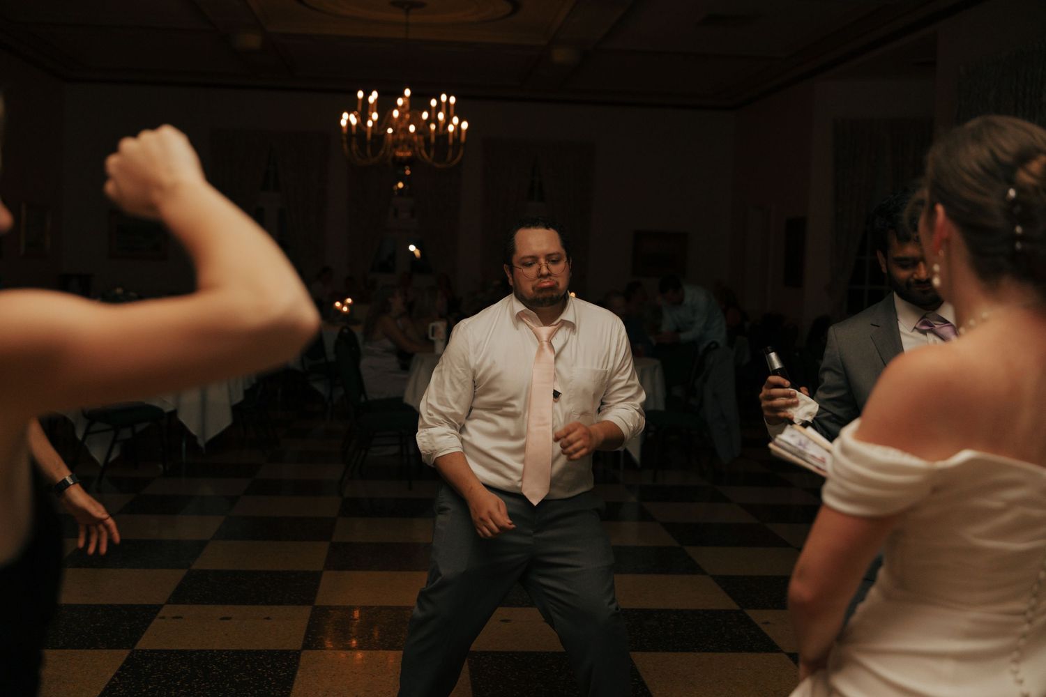 Man dancing energetically at a wedding reception on a black and white checkered dance floor under a chandelier.