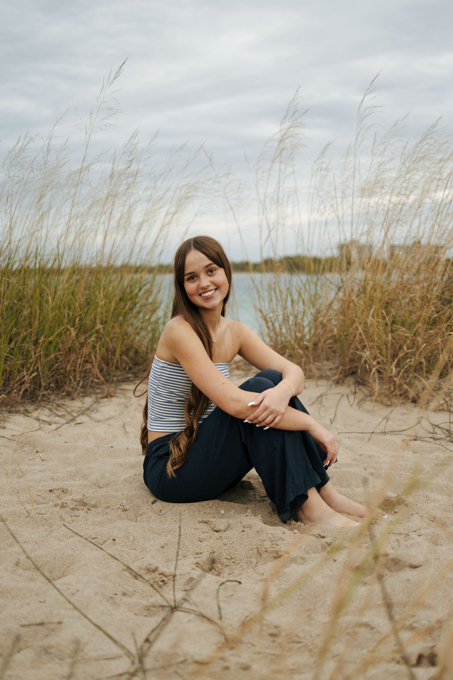Smiling young woman sitting barefoot on sandy beach dunes with tall grass and water in background.
