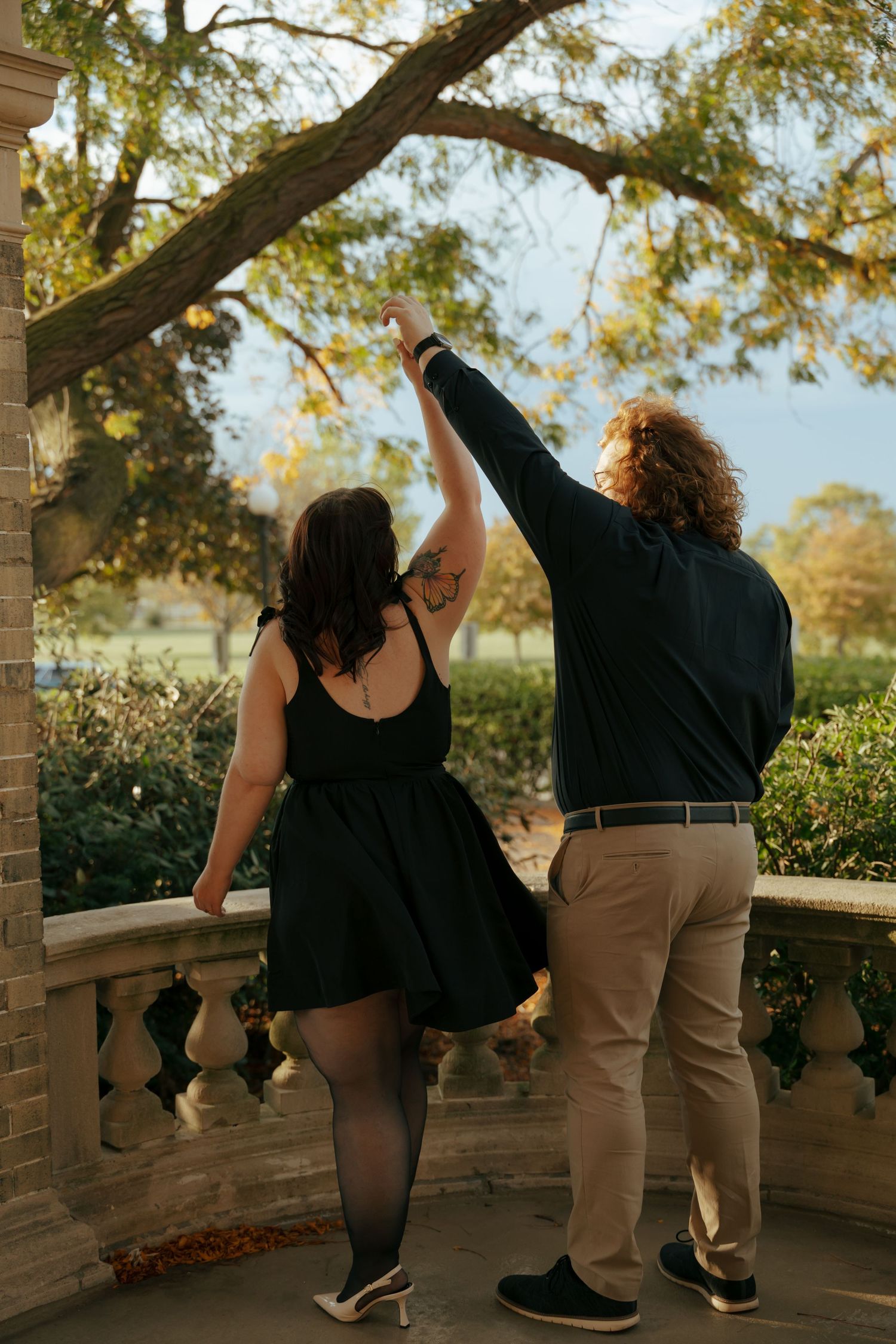 Couple raising joined hands on a stone balcony surrounded by autumn trees.