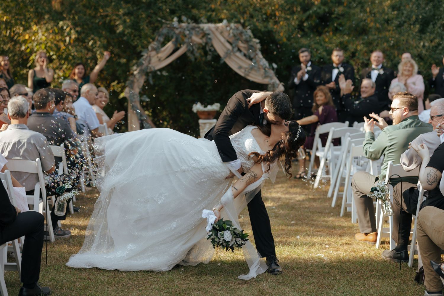 Groom dips his bride for a kiss at their outdoor wedding ceremony as guests cheer and applaud.