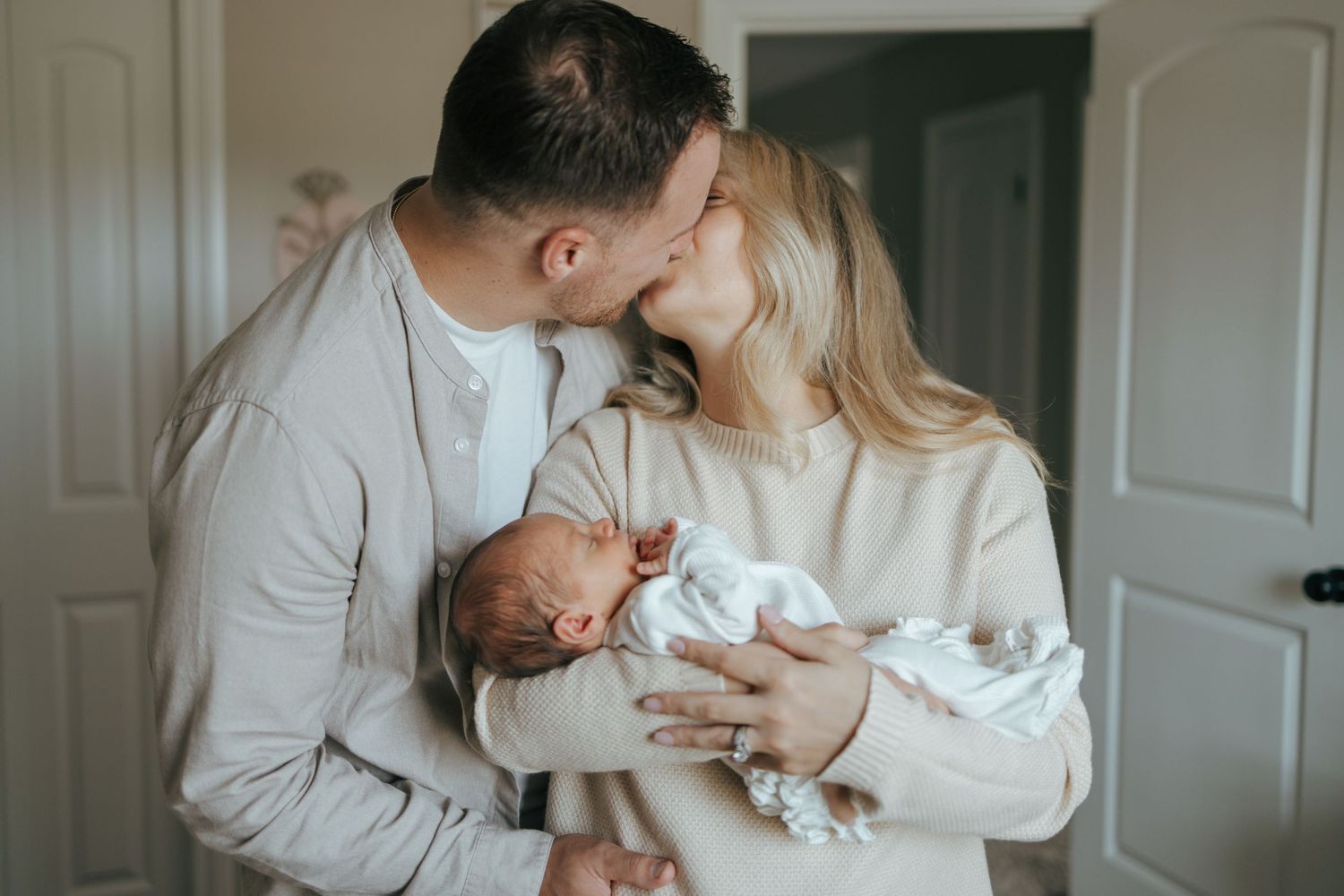 New parents sharing a kiss while tenderly holding their newborn baby together at home.