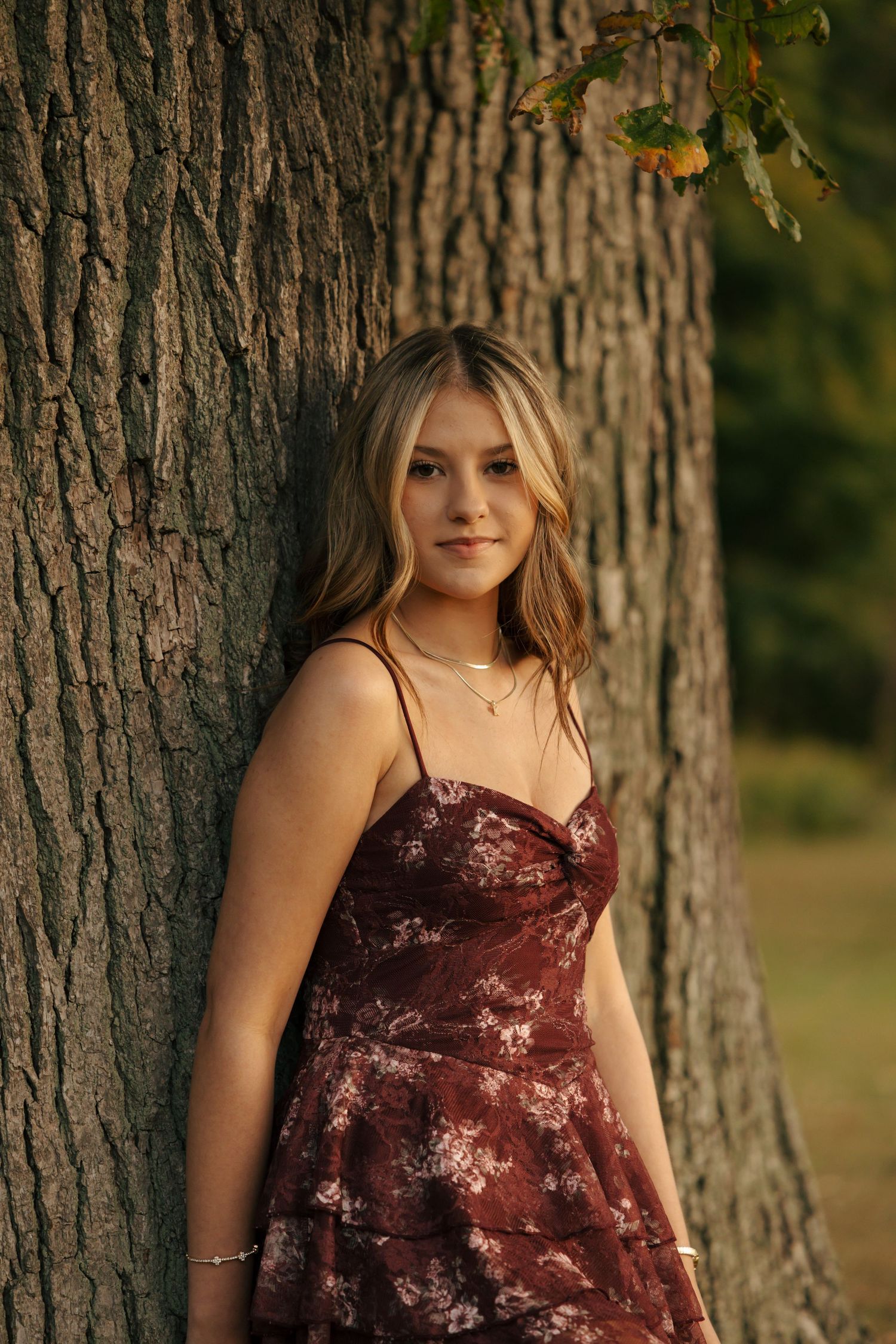 Young woman in burgundy floral dress leaning against a large tree trunk outdoors.