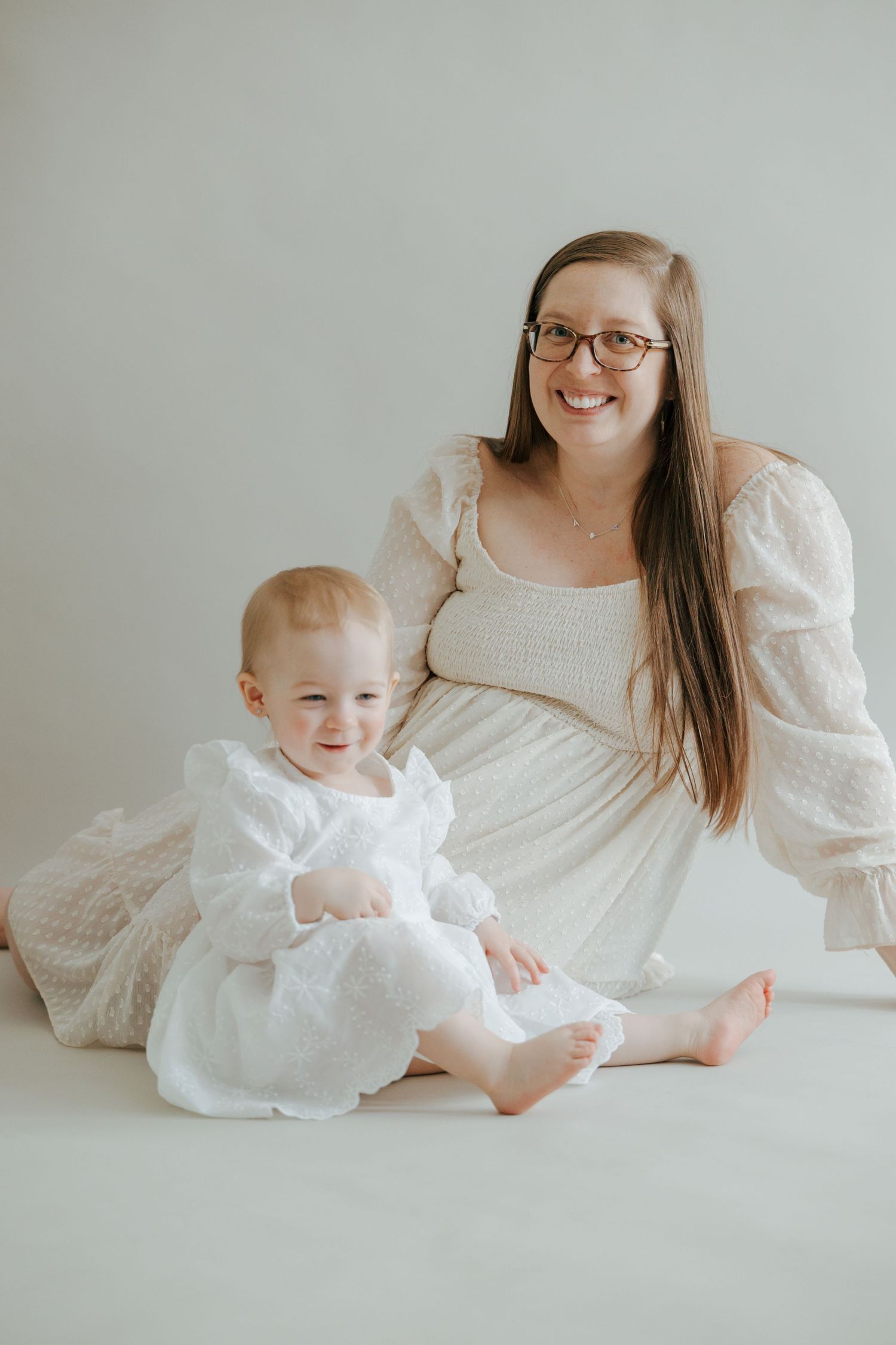 Smiling pregnant woman in cream dress sitting with happy baby girl in white dress.