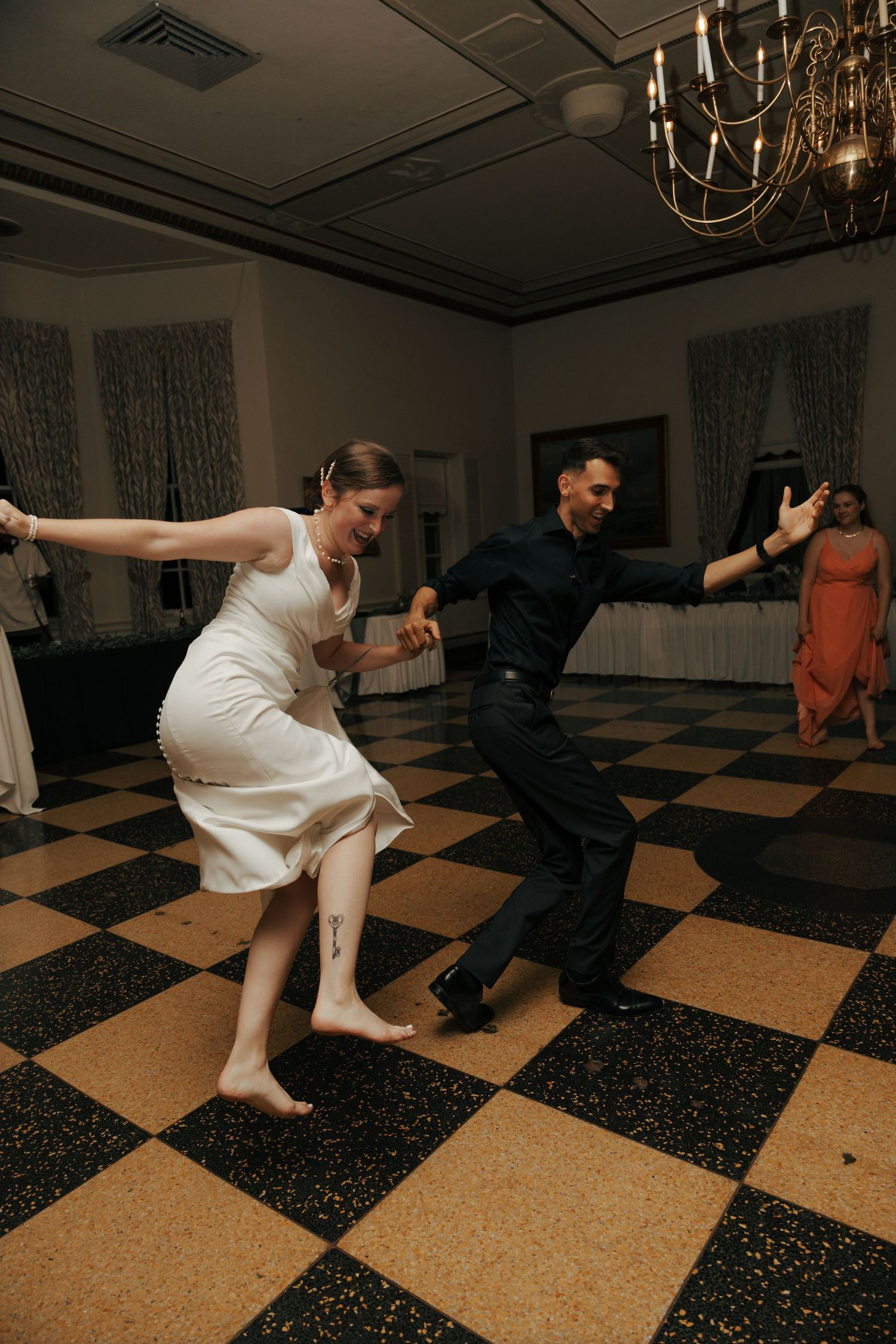 Barefoot bride and groom performing energetic dance on checkered wedding reception floor.