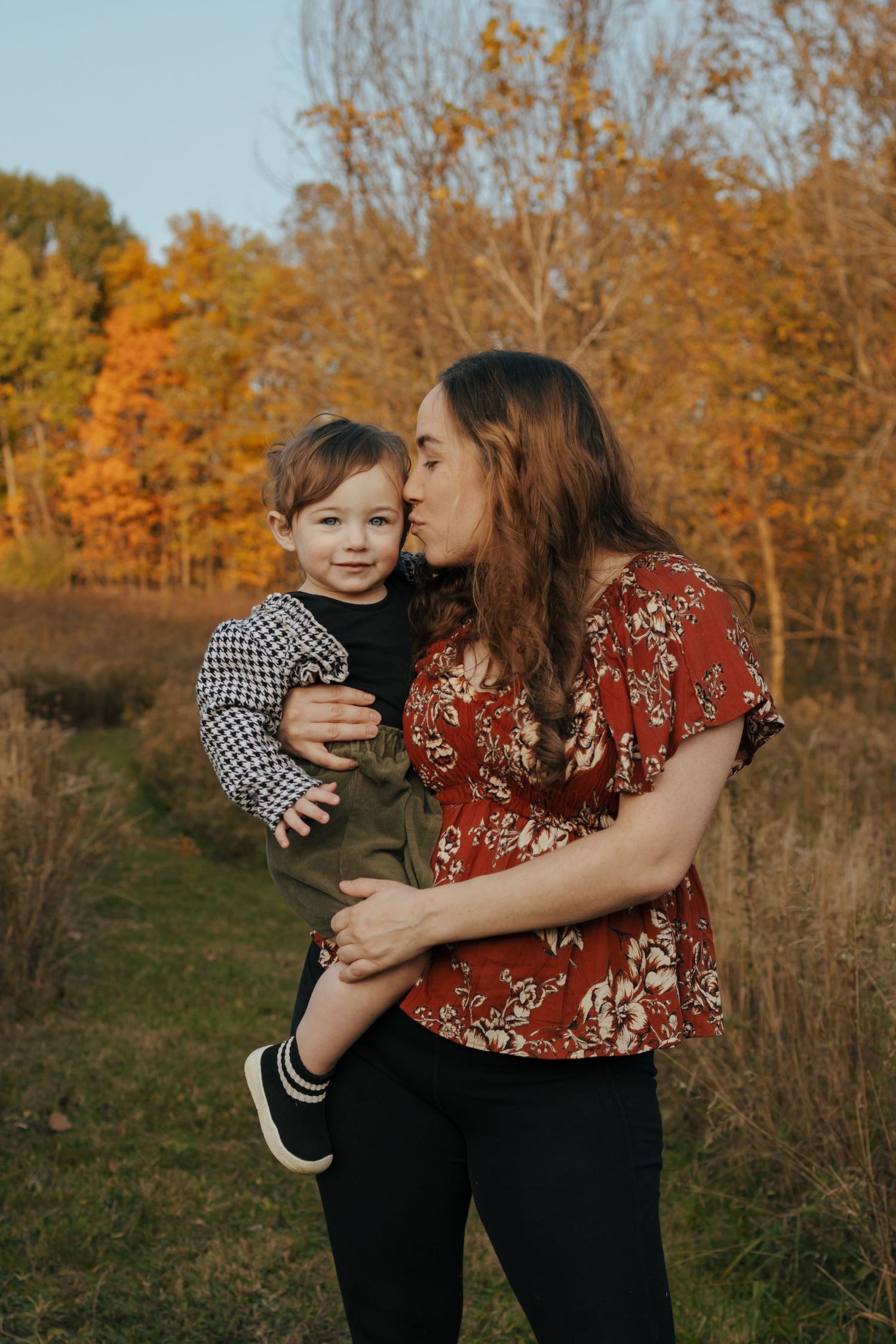 A mother tenderly kisses her toddler son while holding him in an autumn outdoor setting.