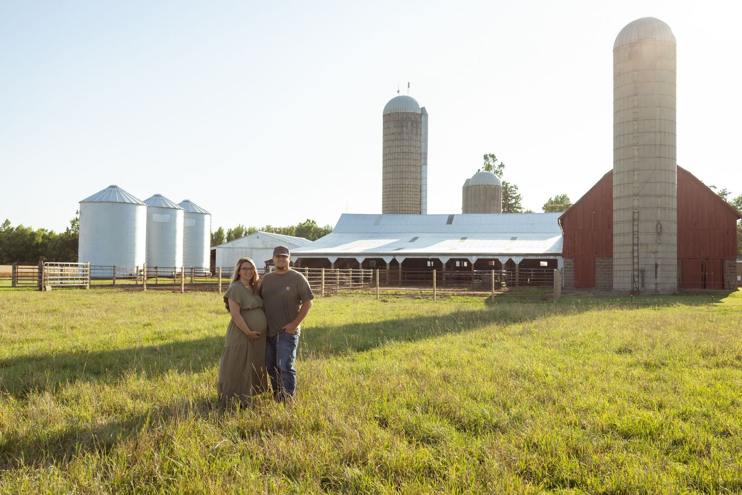 Couple standing together in a lush green farm field with red barn and grain silos in background.