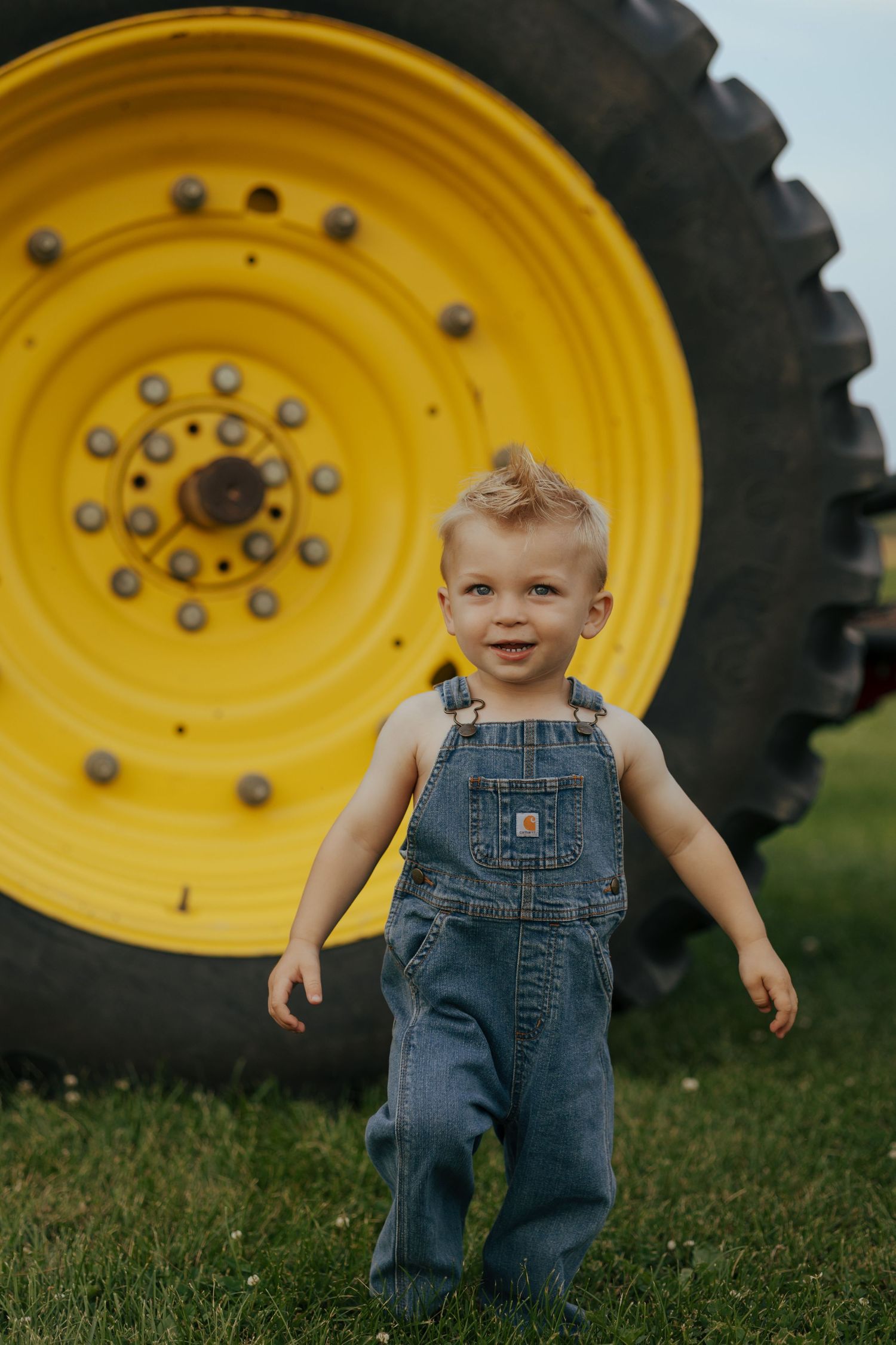Happy toddler in denim overalls standing in front of a large yellow tractor wheel on green grass.