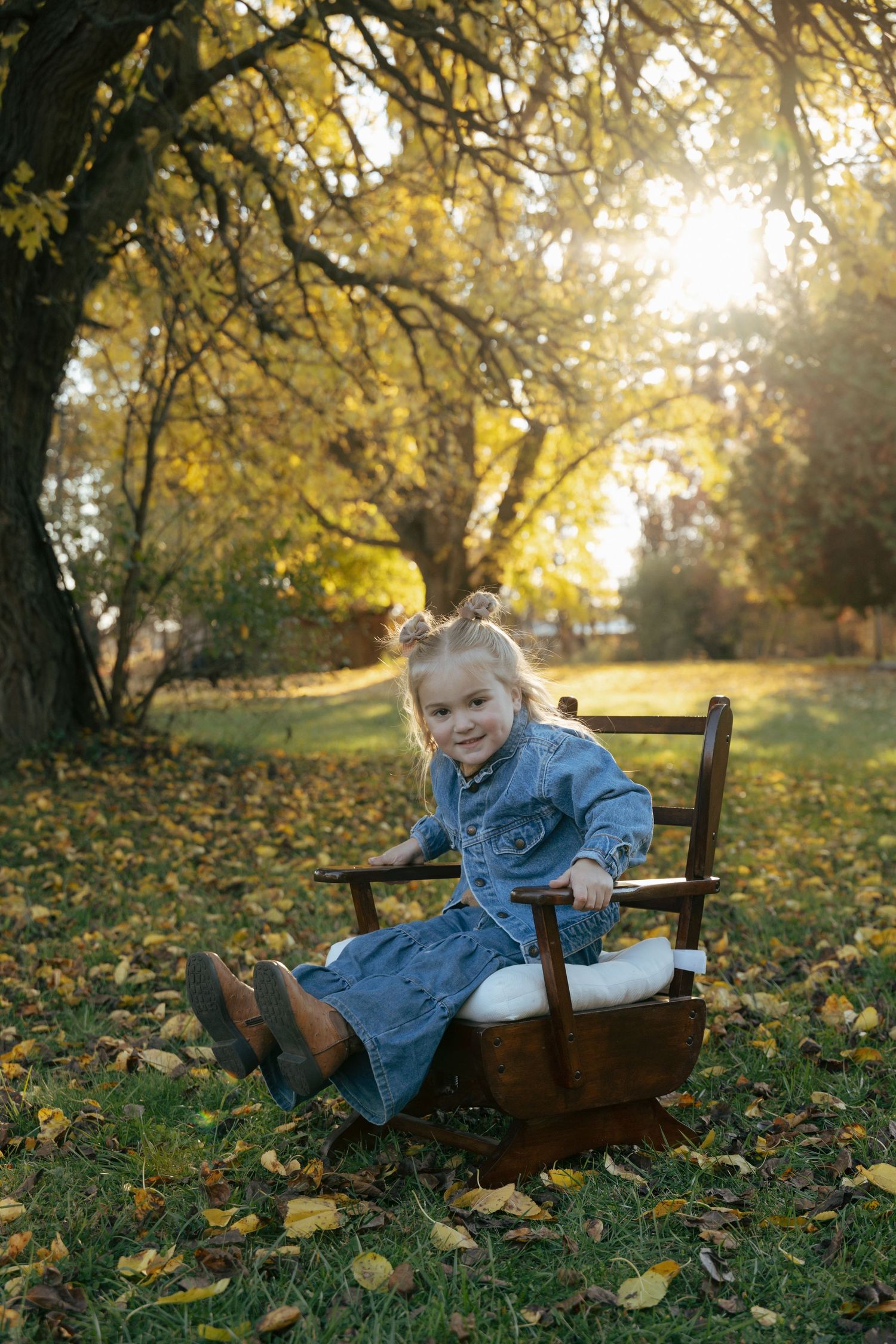 Adorable little girl in denim dress sitting in wooden chair surrounded by fallen autumn leaves in park.