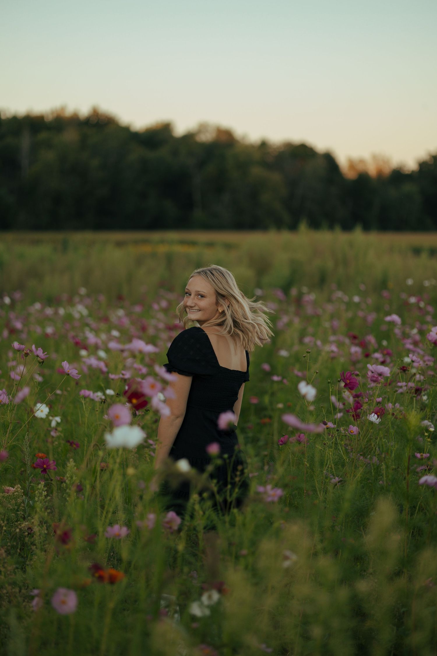 Blonde teenage girl in black dress standing in pink wildflower field at golden hour sunset.