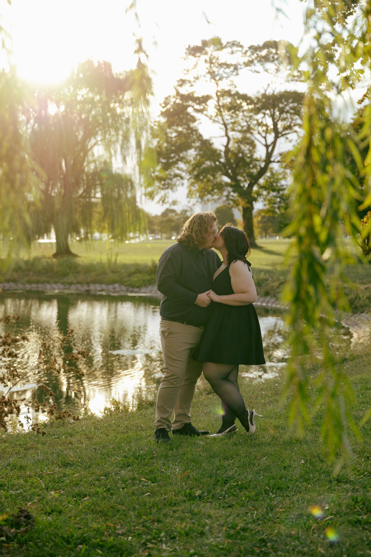 Couple sharing a kiss near a sunlit pond framed by weeping willow branches.