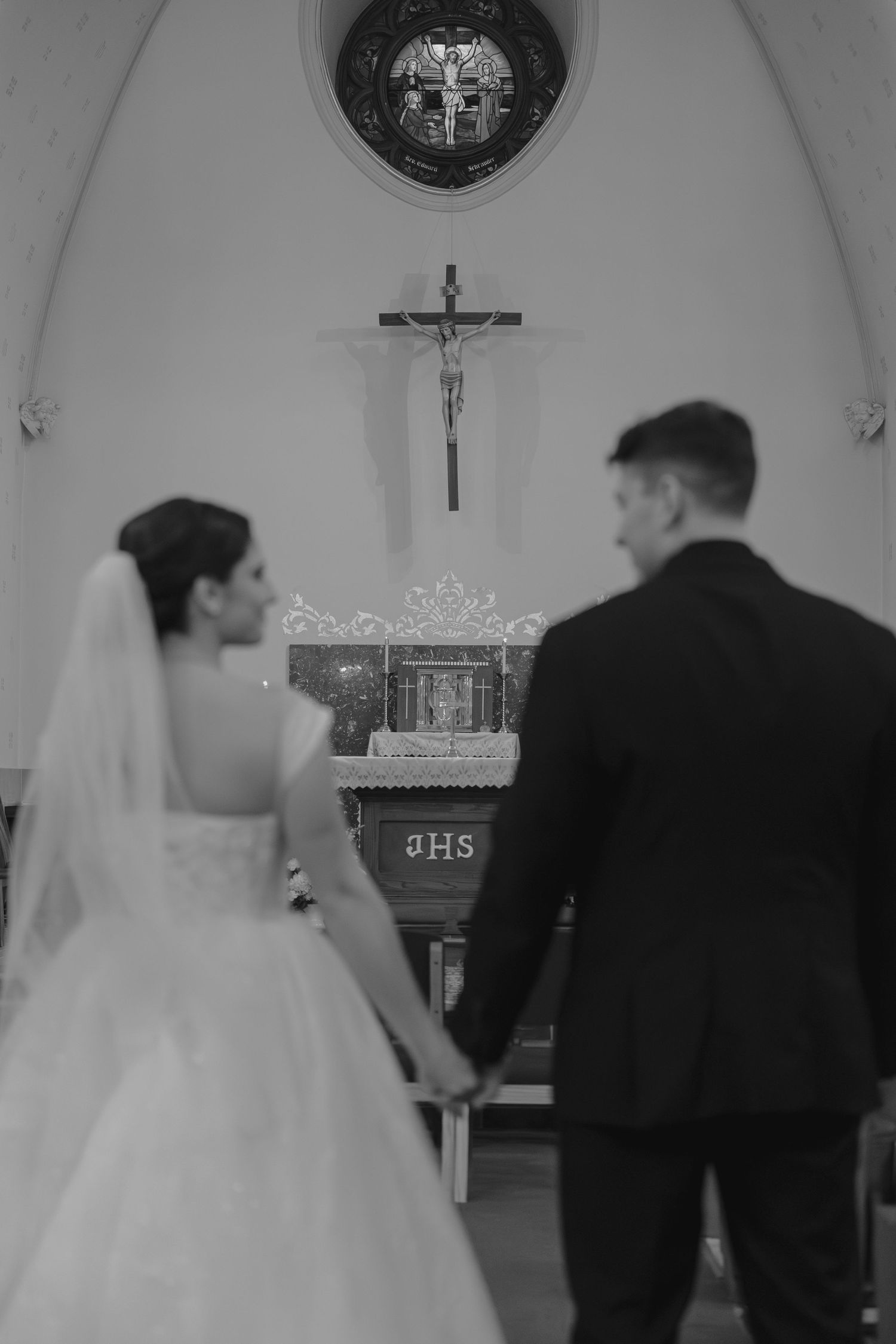 Bride and groom holding hands facing church altar with crucifix in black and white wedding photo.