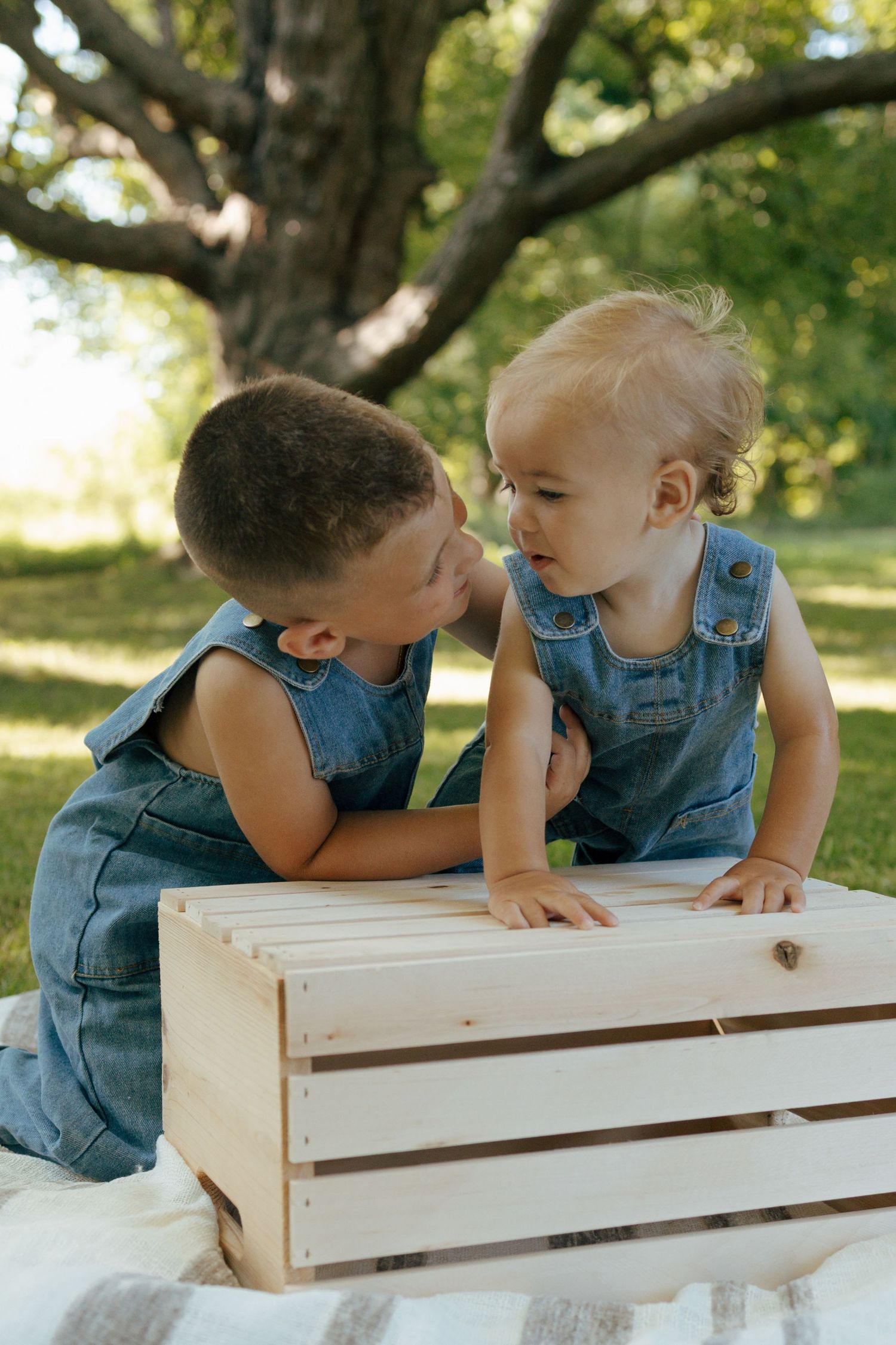 Two toddlers in matching denim overalls interact curiously over a wooden crate in a park.
