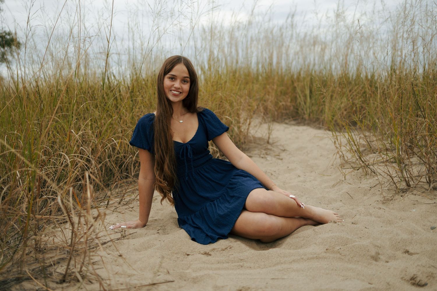 Young woman in navy blue dress sitting on sandy beach path surrounded by tall golden grass.