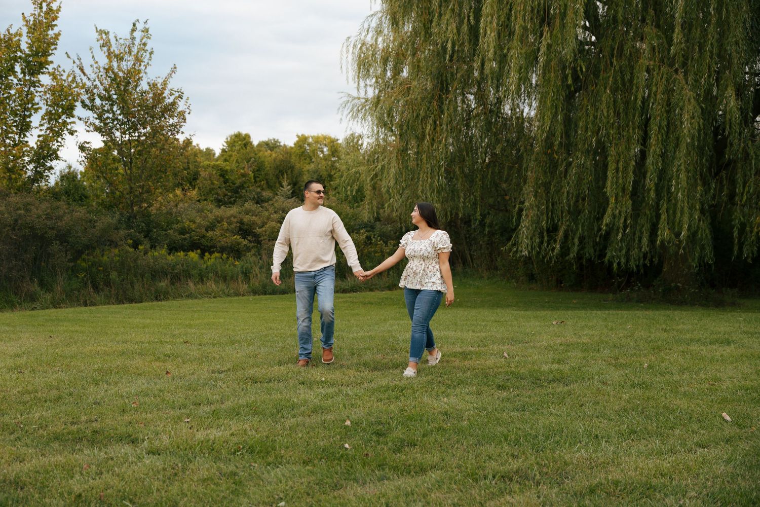 Happy couple holding hands and running together through a lush green park near a willow tree.