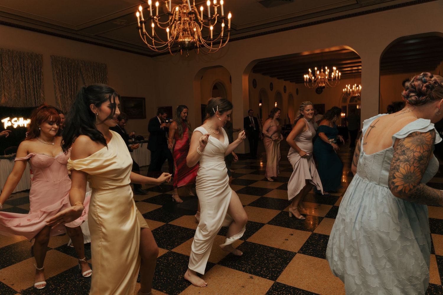 Wedding guests in formal attire dancing together on a checkered ballroom floor under a chandelier.