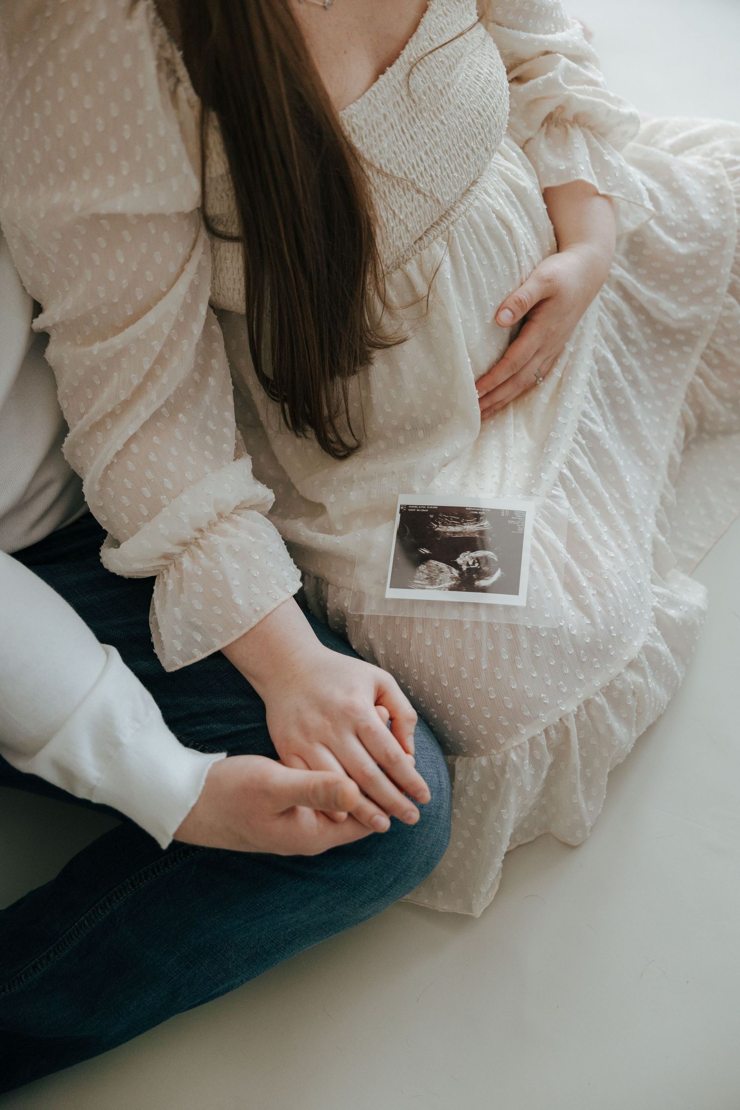 Couple sitting together holding hands with ultrasound photo resting on pregnant belly.