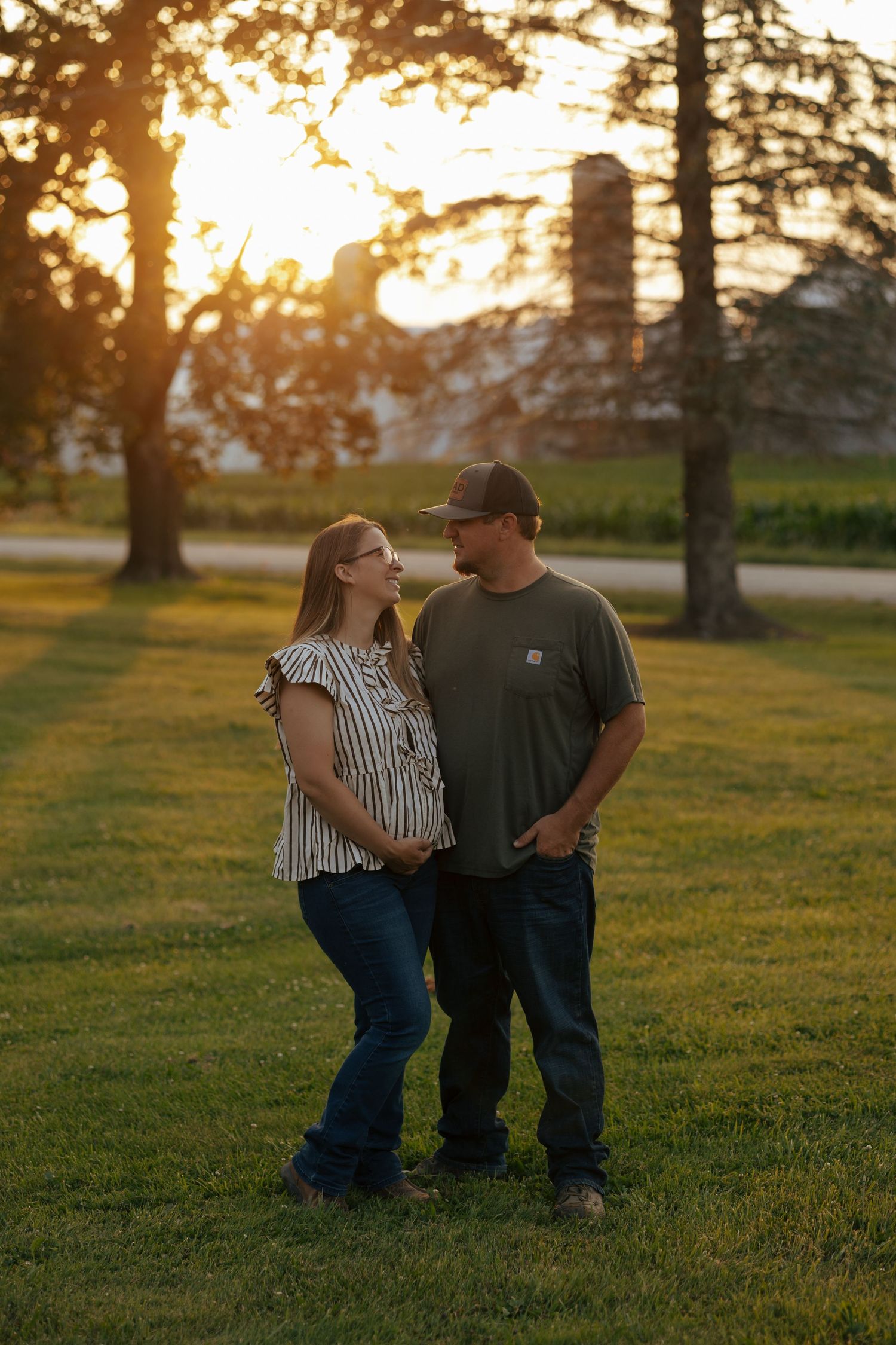 Couple sharing a loving glance at golden hour in a green field with trees and silo behind them.